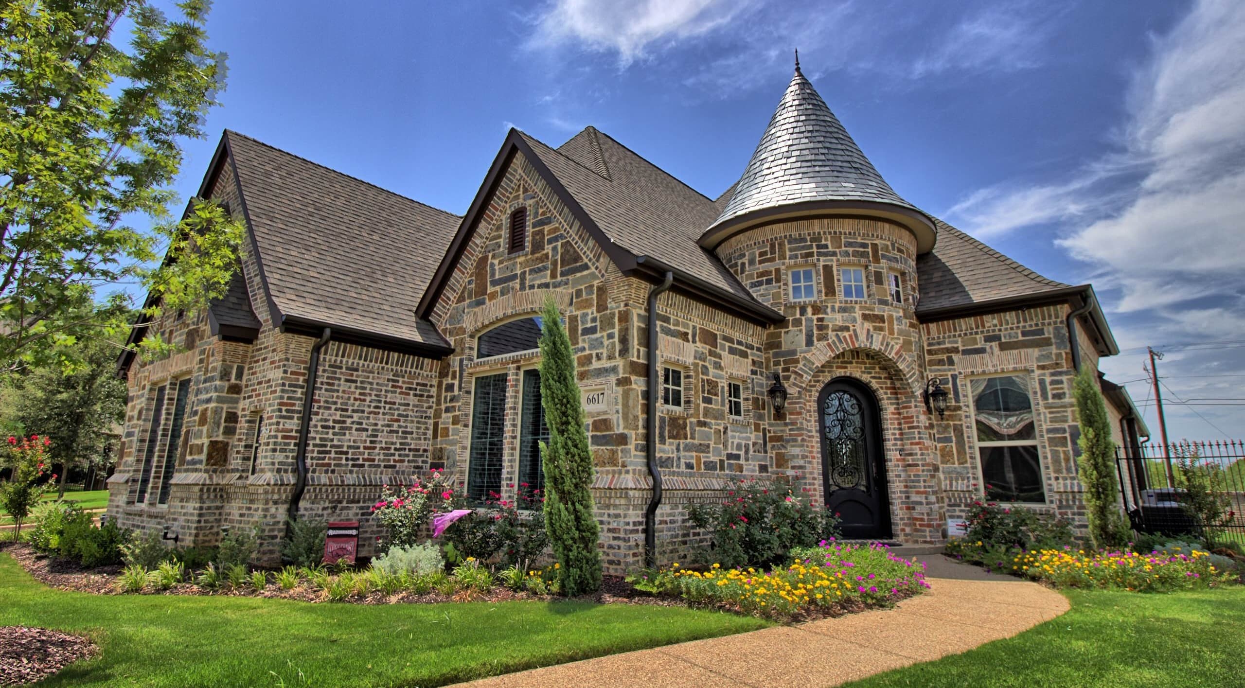 Stone and brick facade with a round turret under a conical gray shingle roof, arched iron-and-glass front door, and flower-lined curved walkway