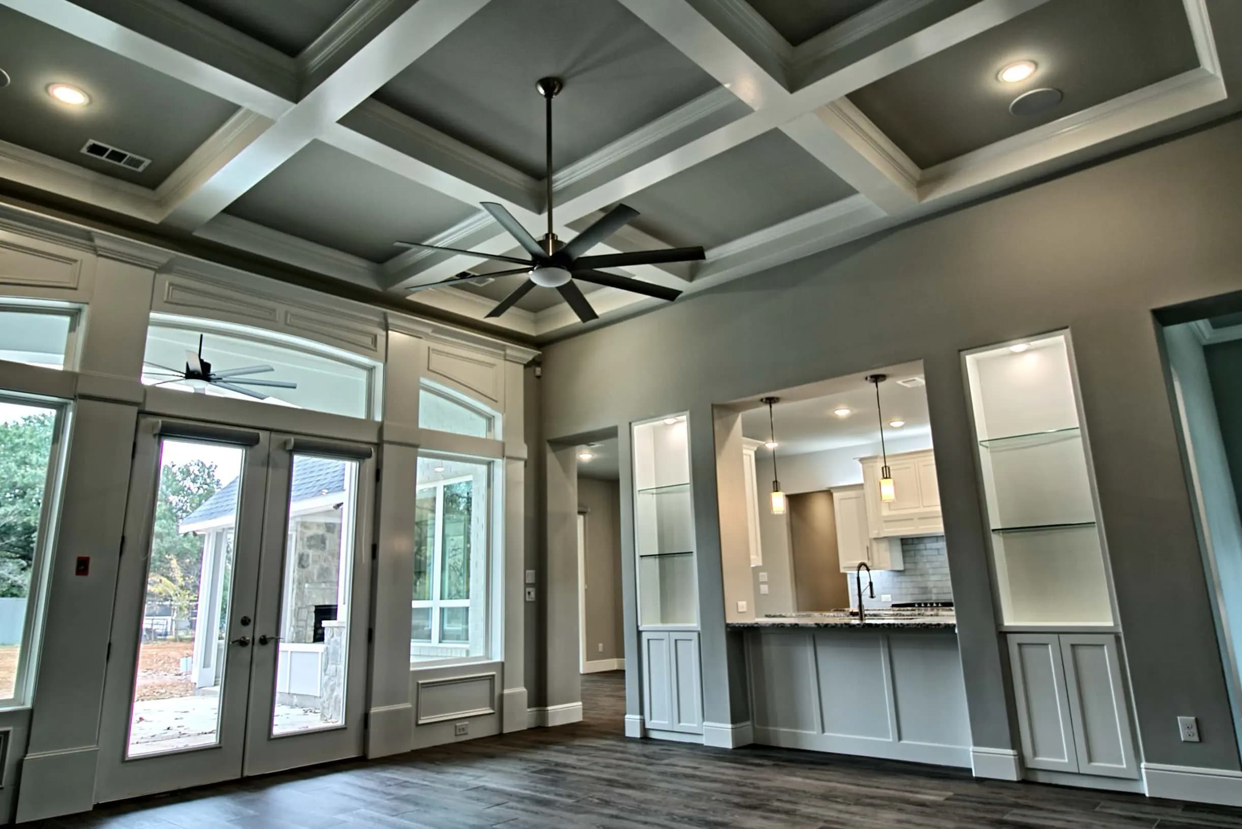 Living room with coffered ceiling, large ceiling fan, glass-front built-ins, hardwood floors and French doors to a stone patio