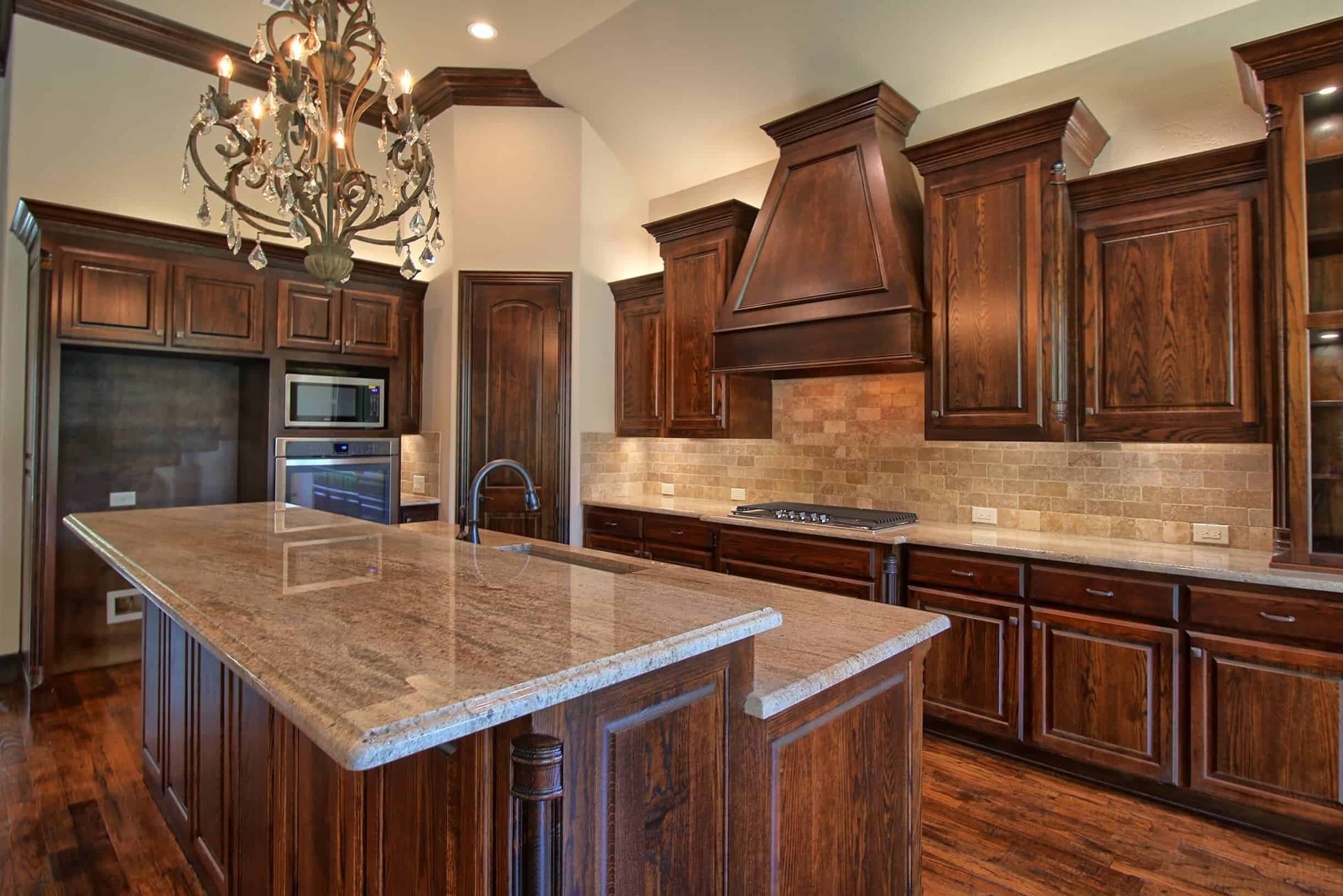 Kitchen with dark stained wood cabinetry and hood, travertine backsplash, large granite island and crystal chandelier over wood floors