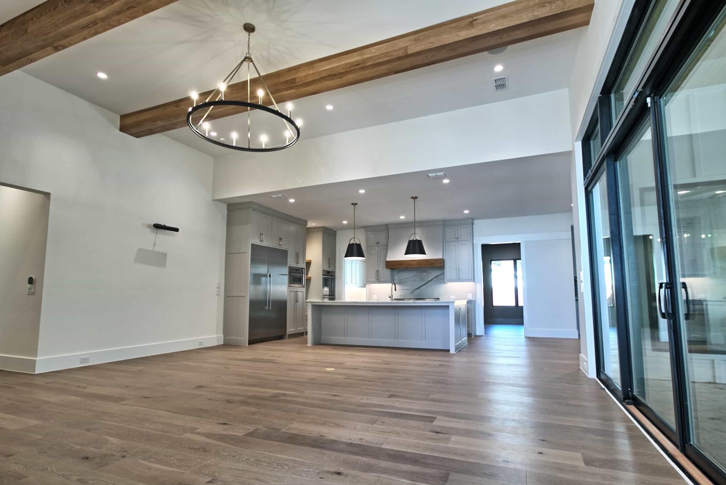 Great room with wood ceiling beams, black ring chandelier and wide-plank floors, open to a gray shaker kitchen with island, black pendants and black-framed sliding doors