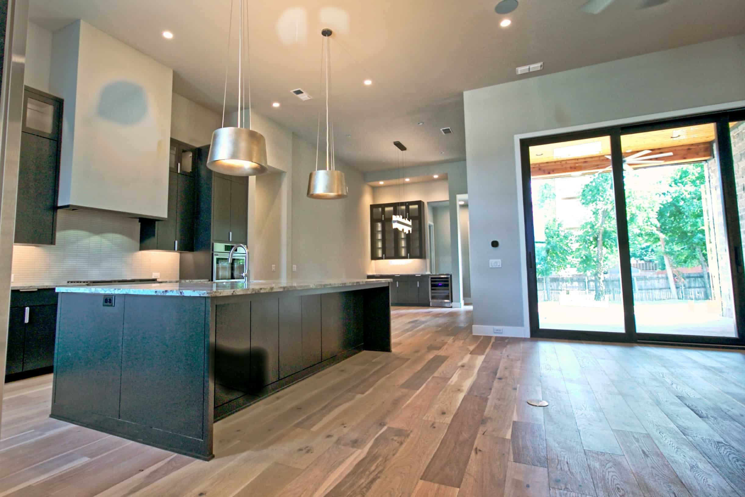 Open kitchen with charcoal flat-panel cabinetry, long granite-topped island, silver drum pendants, wide-plank wood floors and black-framed sliding doors to a covered patio