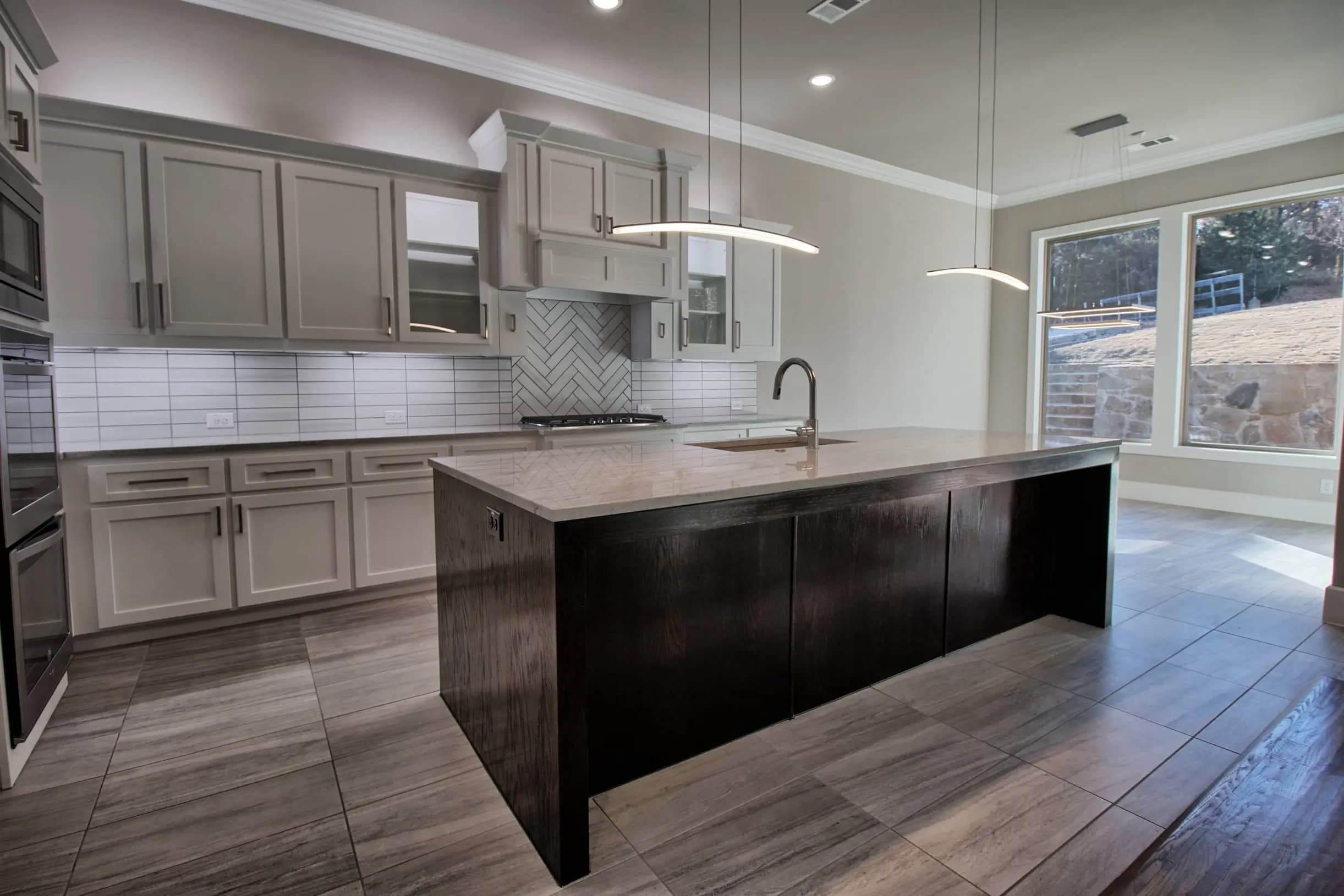 Kitchen with light gray shaker cabinets, herringbone tile backsplash, large picture windows, dark wood island with quartz top and sink, pendant lights and large-format tile floors