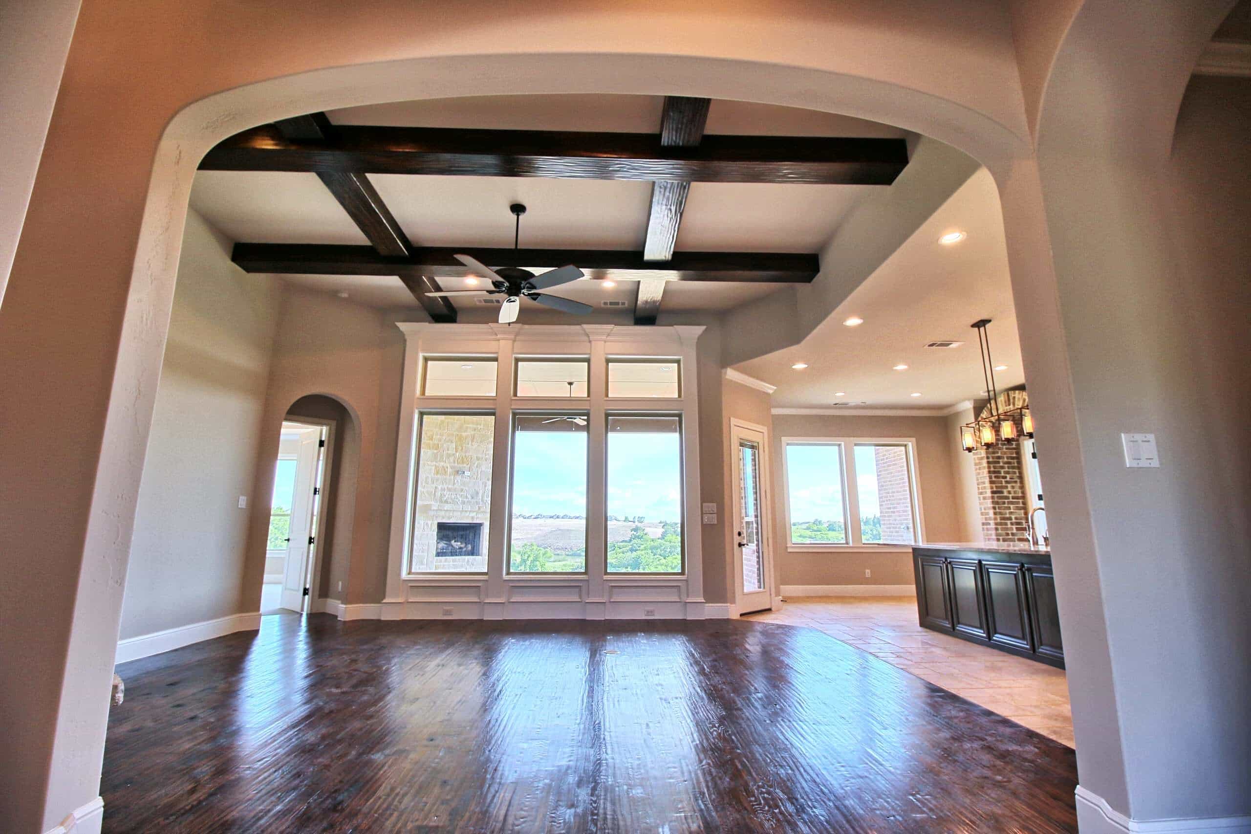 Living room seen through a wide arch, with dark wood ceiling beams, hand-scraped hardwood floors, tall windows and an open kitchen beyond