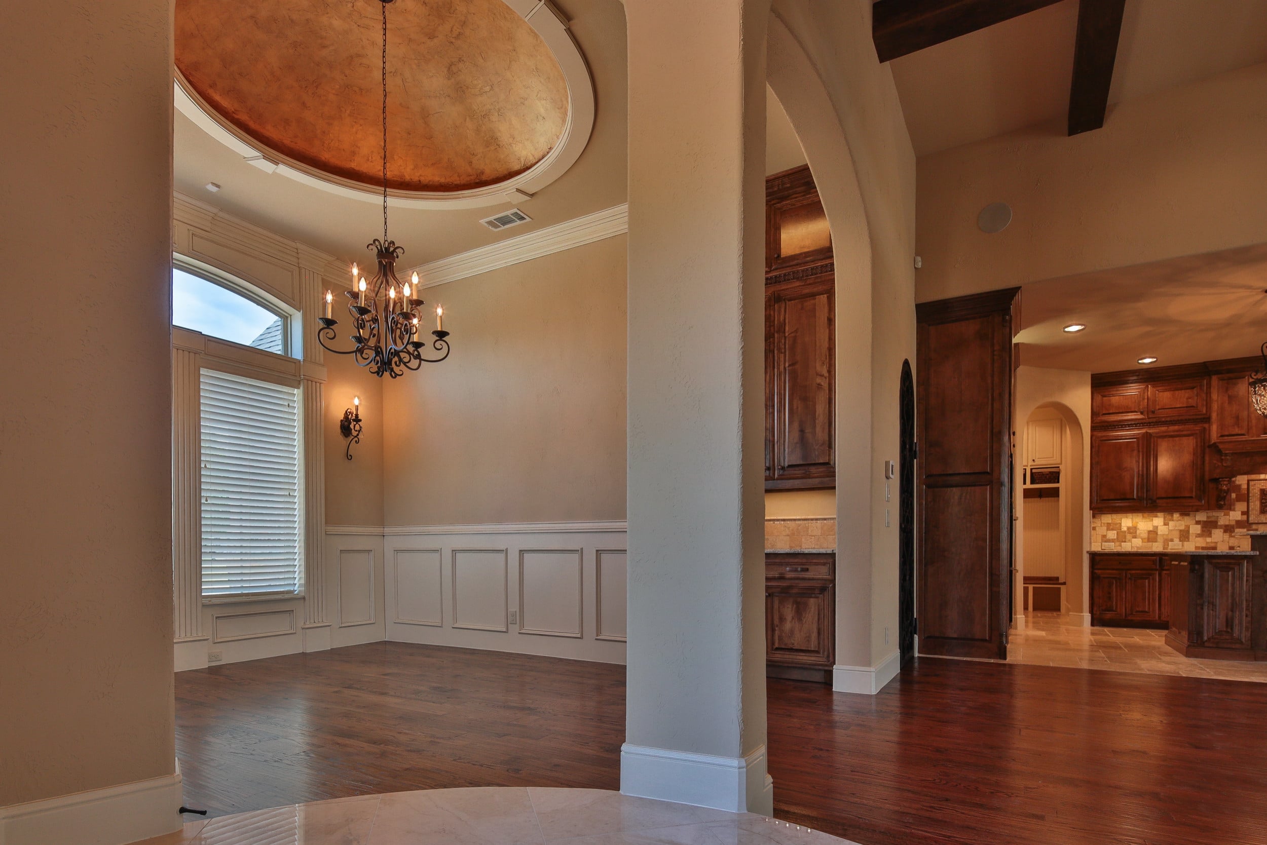 Formal dining room with copper-toned domed ceiling, wrought iron chandelier, white wainscoting, and hardwood floor, beside an archway to the kitchen