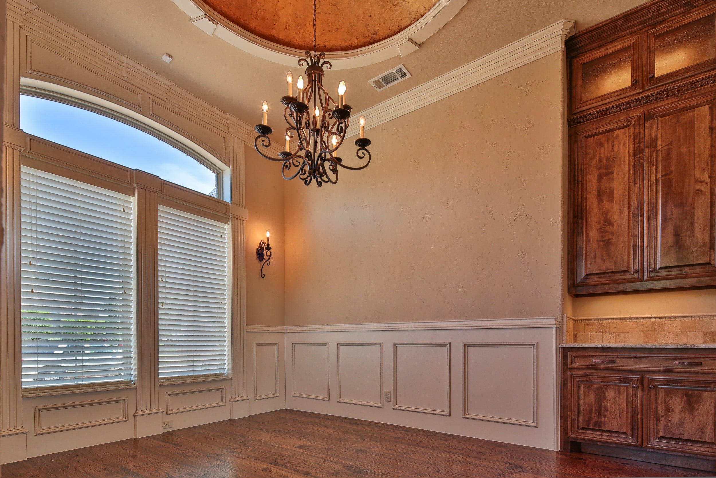 Dining room with tall windows under an arched transom, wrought iron chandelier, white wainscoting, hardwood floor, and stained wood built-in buffet