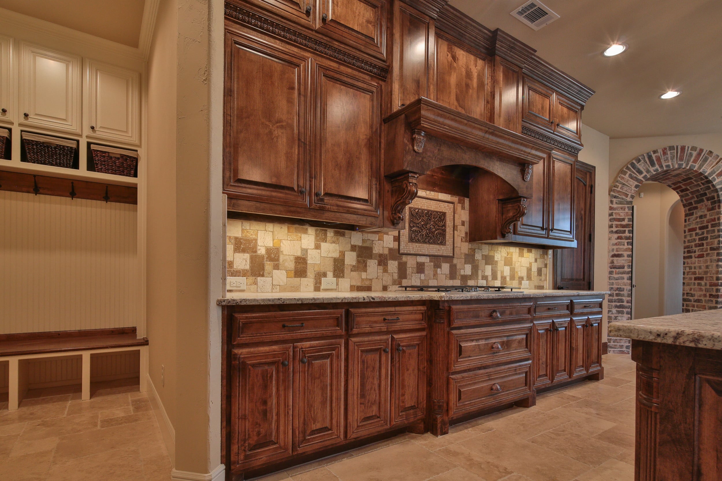 Kitchen cooktop wall with stained wood cabinetry, wood hood with carved corbels, and travertine backsplash, beside a mudroom bench and brick archway