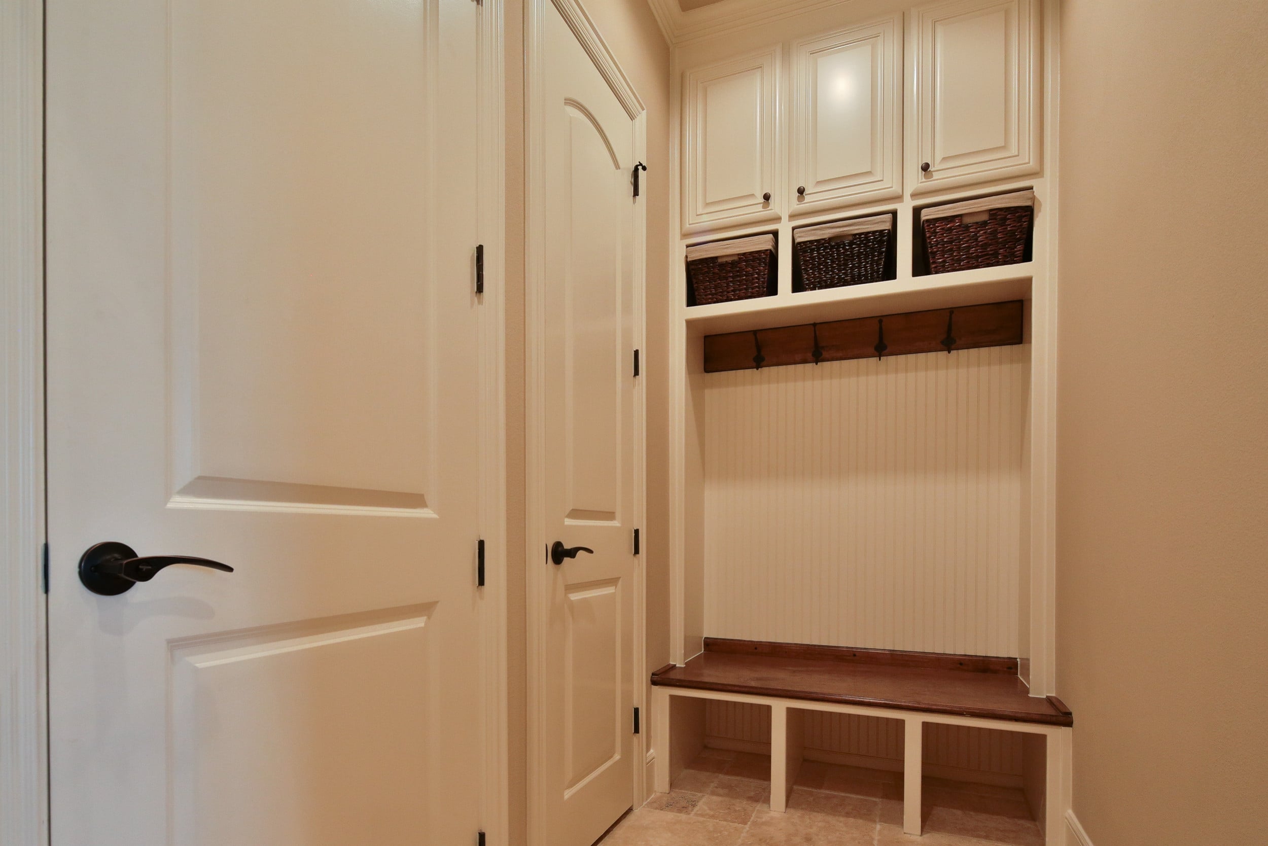 Mudroom drop zone with cream upper cabinets, woven storage baskets, wood coat hook rail, beadboard backing, and stained wood bench over cubbies