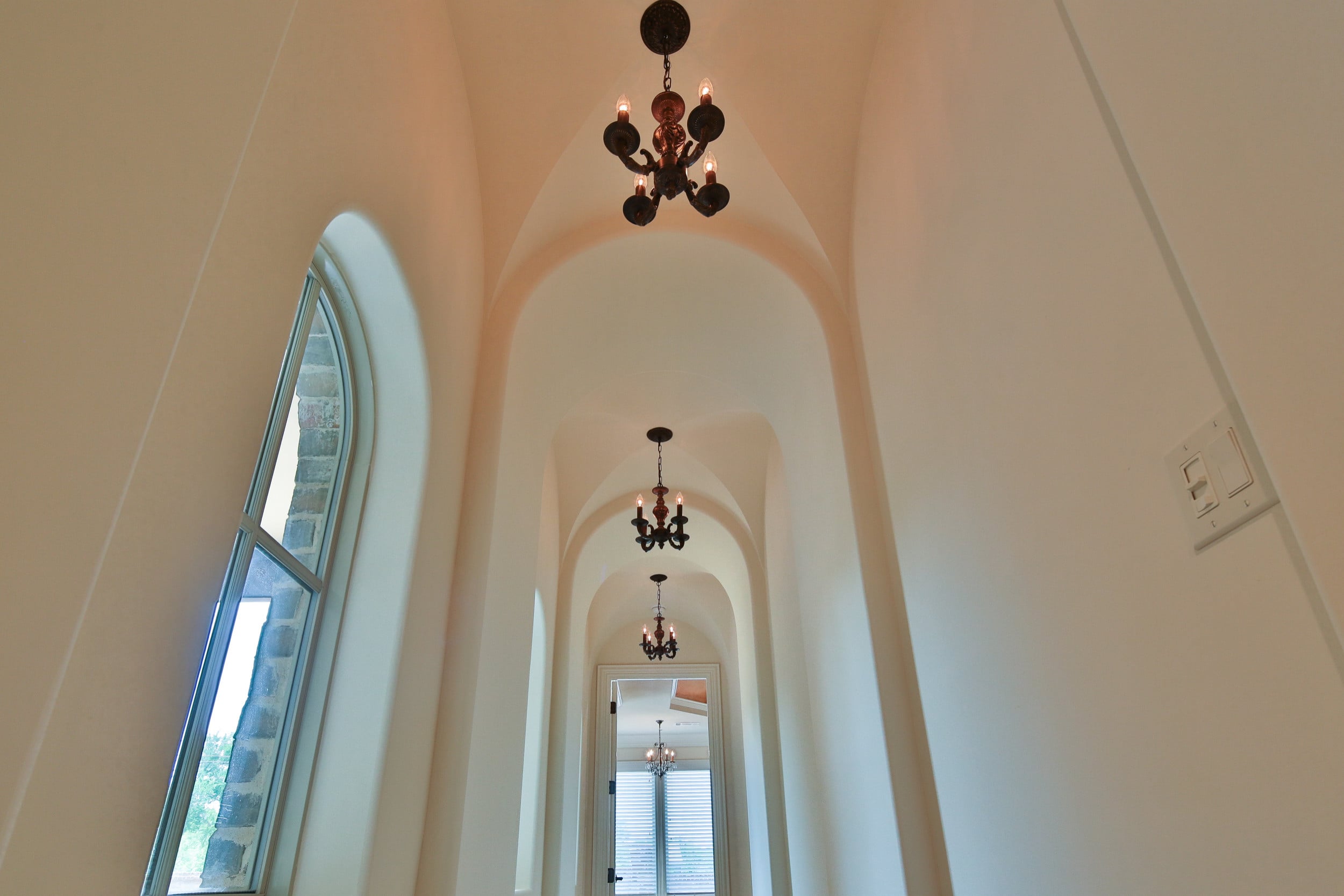 Low-angle view of a groin-vaulted hallway with successive arches, three small bronze candle chandeliers, and a tall arched window