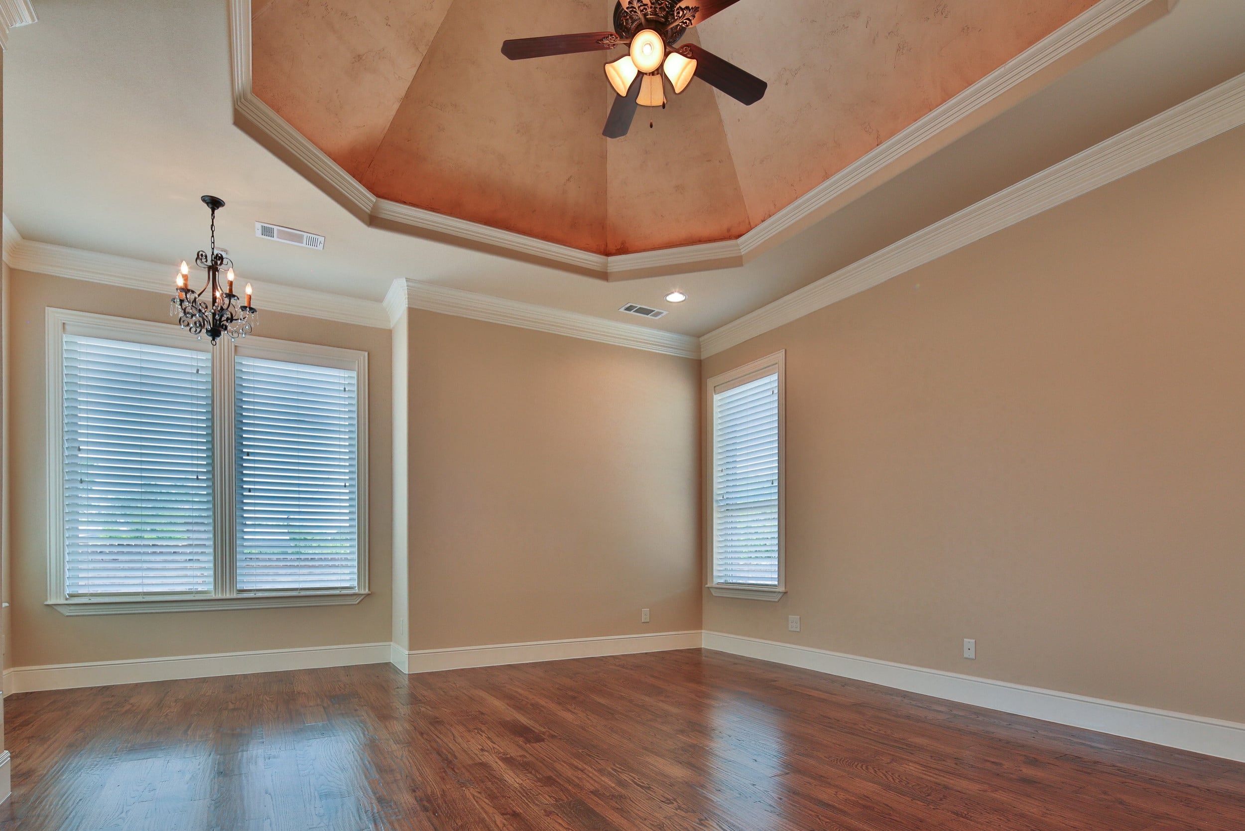 Primary bedroom with copper-finished octagonal tray ceiling, ceiling fan, small crystal chandelier, hardwood floor, crown molding, and windows with white blinds