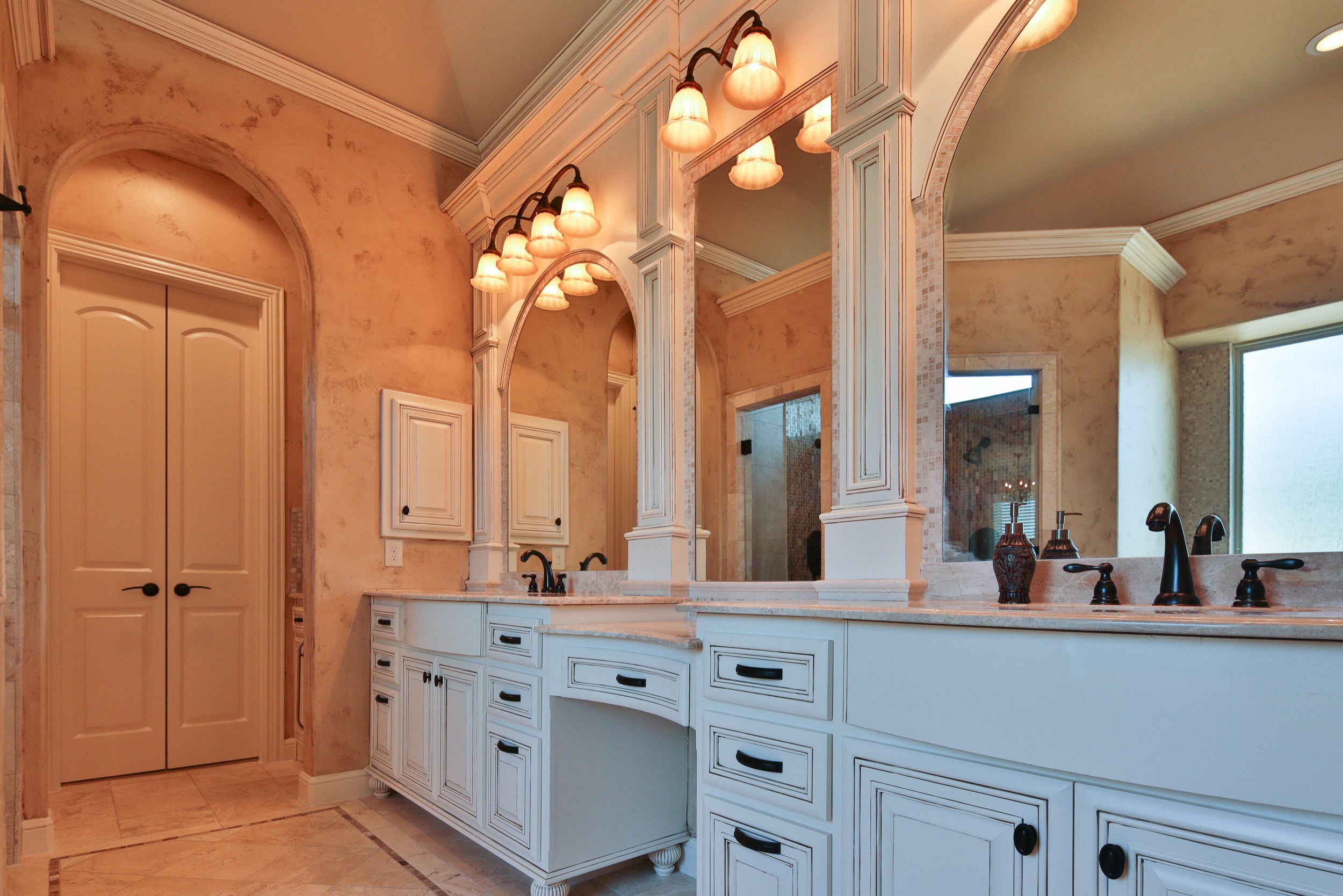 Primary bath with white glazed double vanity, makeup knee space, cream stone counters, oil-rubbed bronze faucets, column-framed mirrors, and glass-shade sconces