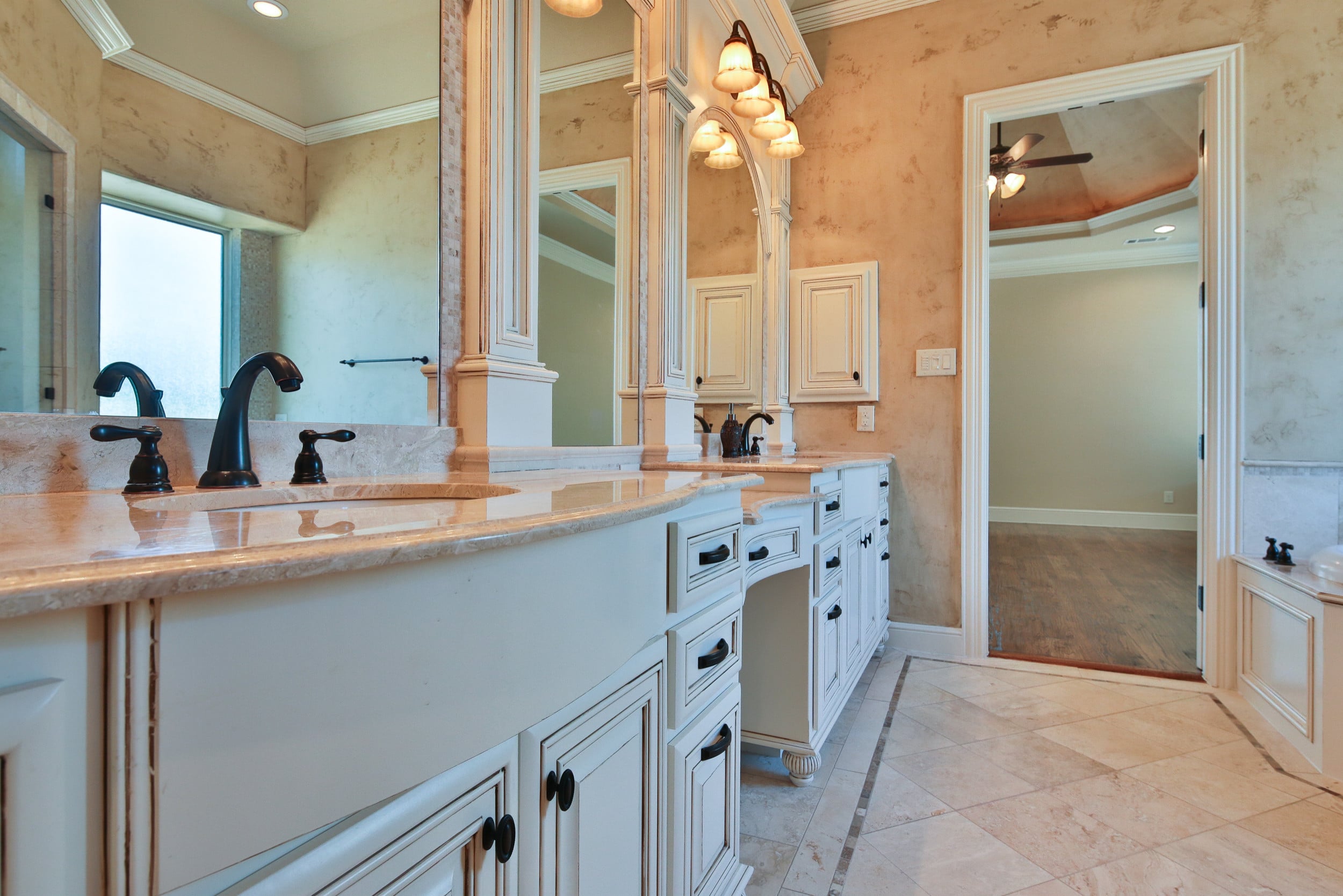 Primary bath with curved white glazed vanity, cream stone counters, oil-rubbed bronze faucets, travertine floor, and an open doorway to the bedroom