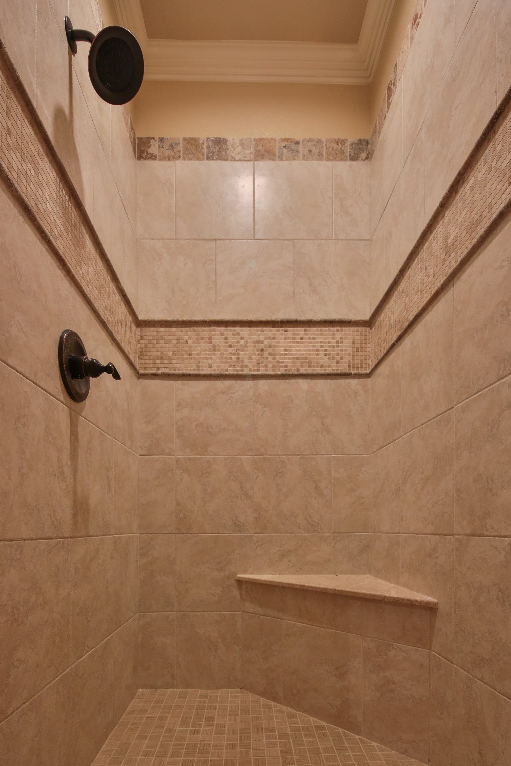 Walk-in shower with beige tile walls, mosaic accent band, tumbled stone top border, oil-rubbed bronze showerhead and valve, and corner seat
