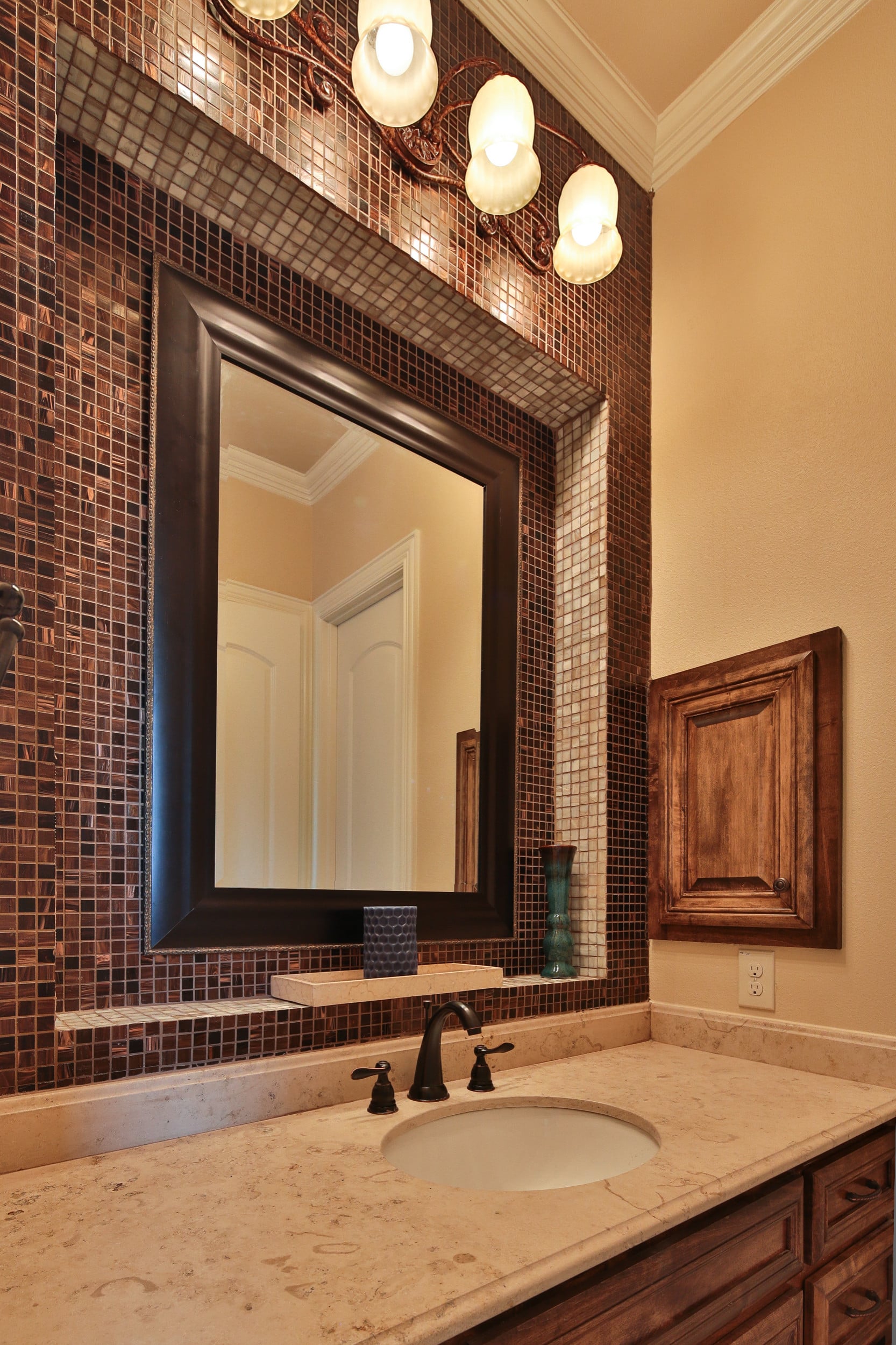 Bathroom vanity with copper-toned glass mosaic accent wall, dark framed mirror, scrolled bronze light fixture, cream stone counter, and oil-rubbed bronze faucet