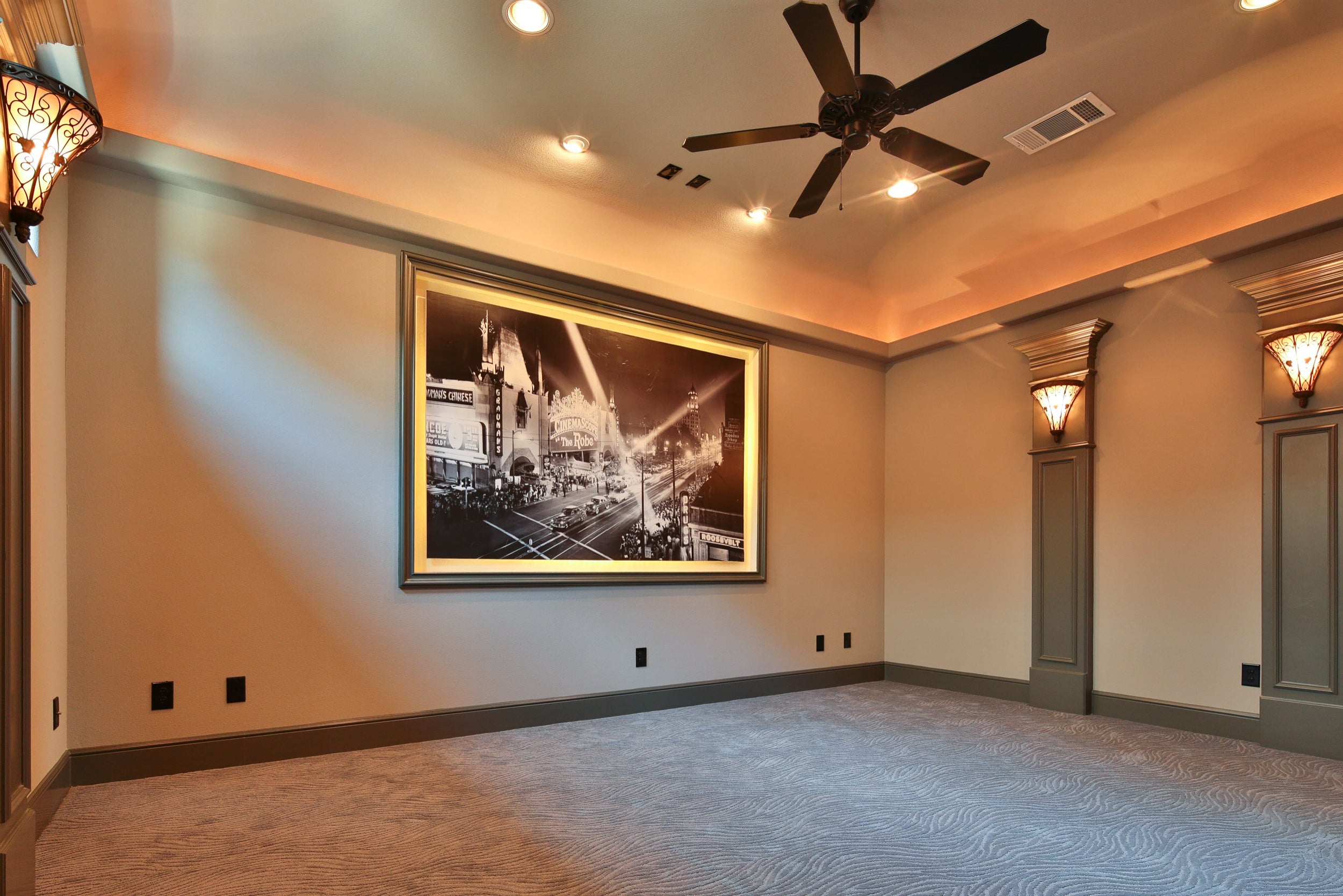 Media room with patterned carpet, cove-lit ceiling, ceiling fan, sconce-topped pilasters, and a framed black-and-white photo of a theater street at night