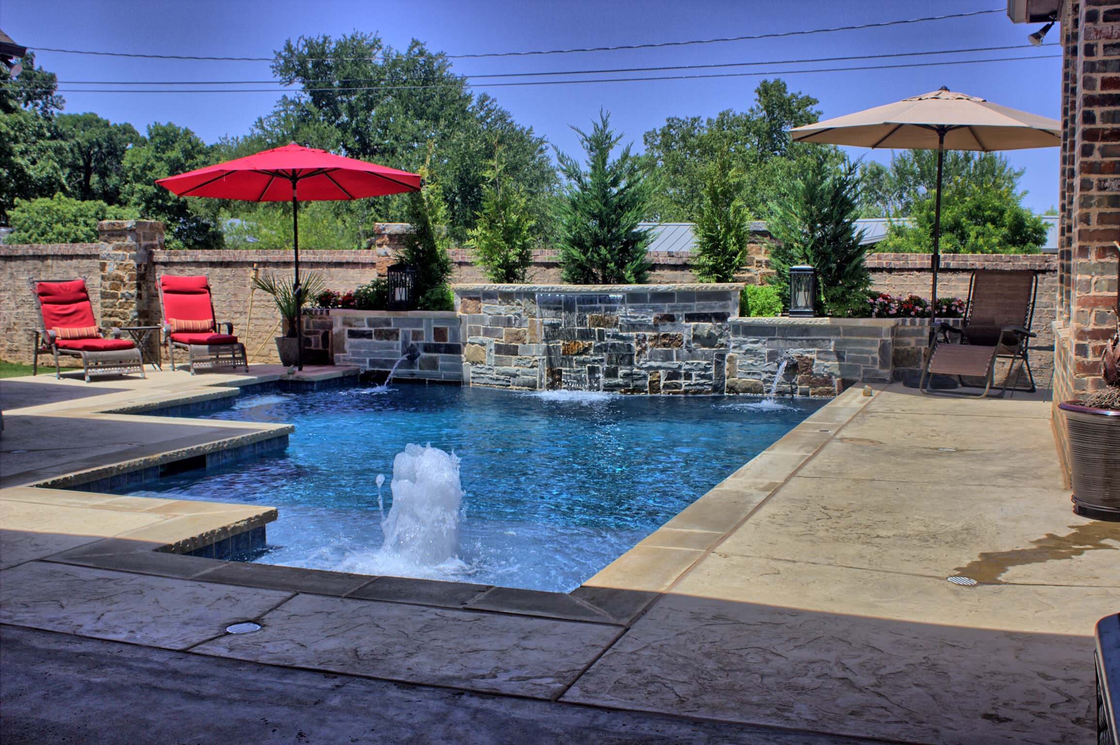 Geometric pool with bubbler, stacked stone water wall and spouts, stamped concrete deck, red lounge chairs and umbrella, and brick perimeter wall