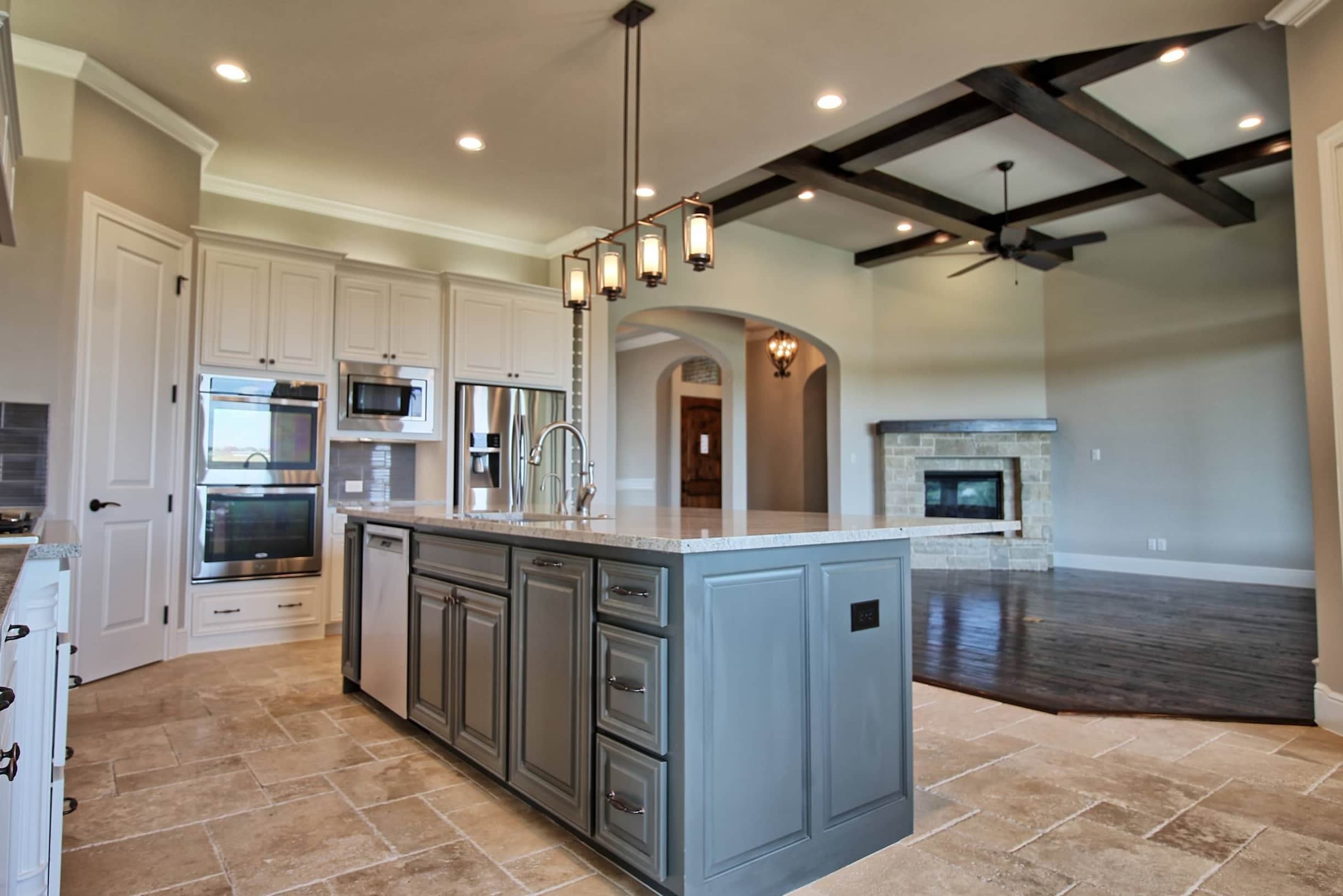 Kitchen with white cabinetry, gray-blue island with granite top, lantern pendants and travertine floor, open to a living room with dark wood ceiling beams and stone fireplace