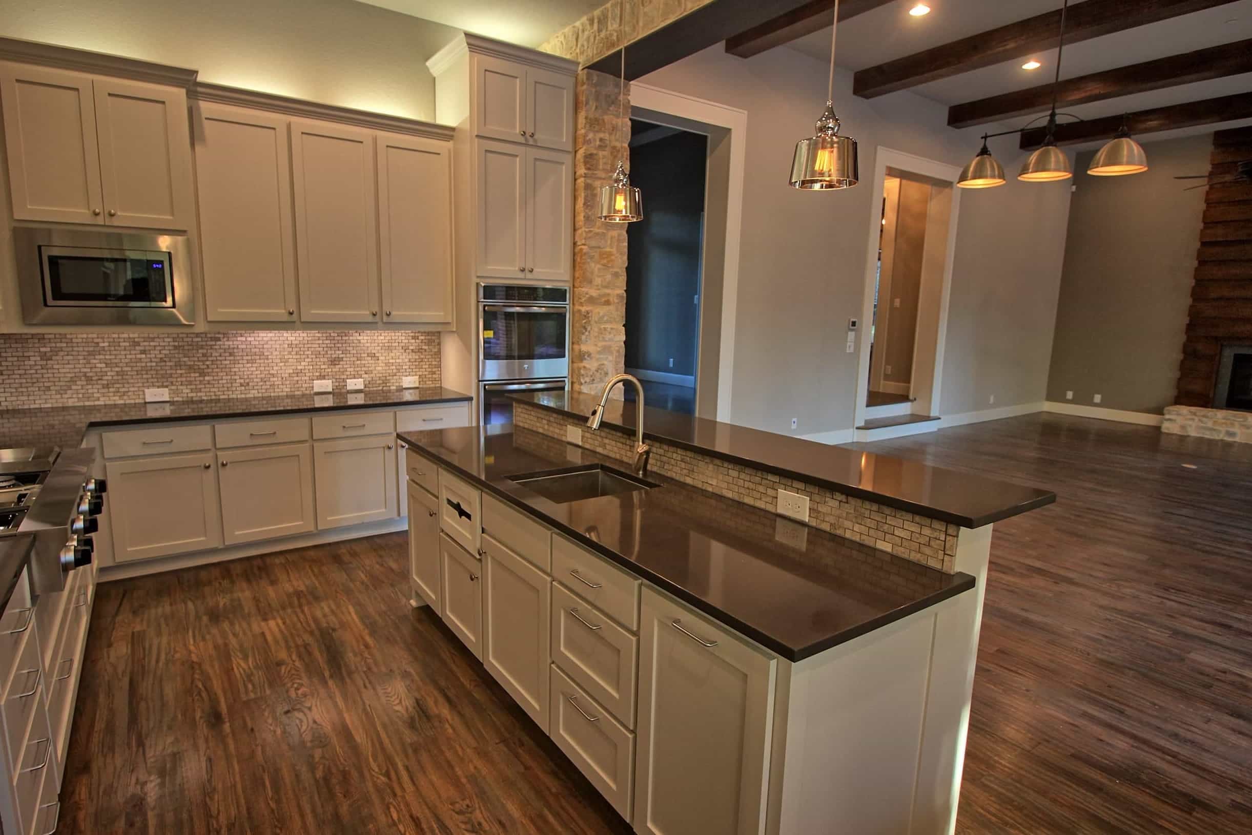 Kitchen with white shaker cabinets, dark quartz countertops, raised island bar with stone tile, stainless wall ovens and pendant lights, open to a beamed living room with wood floors