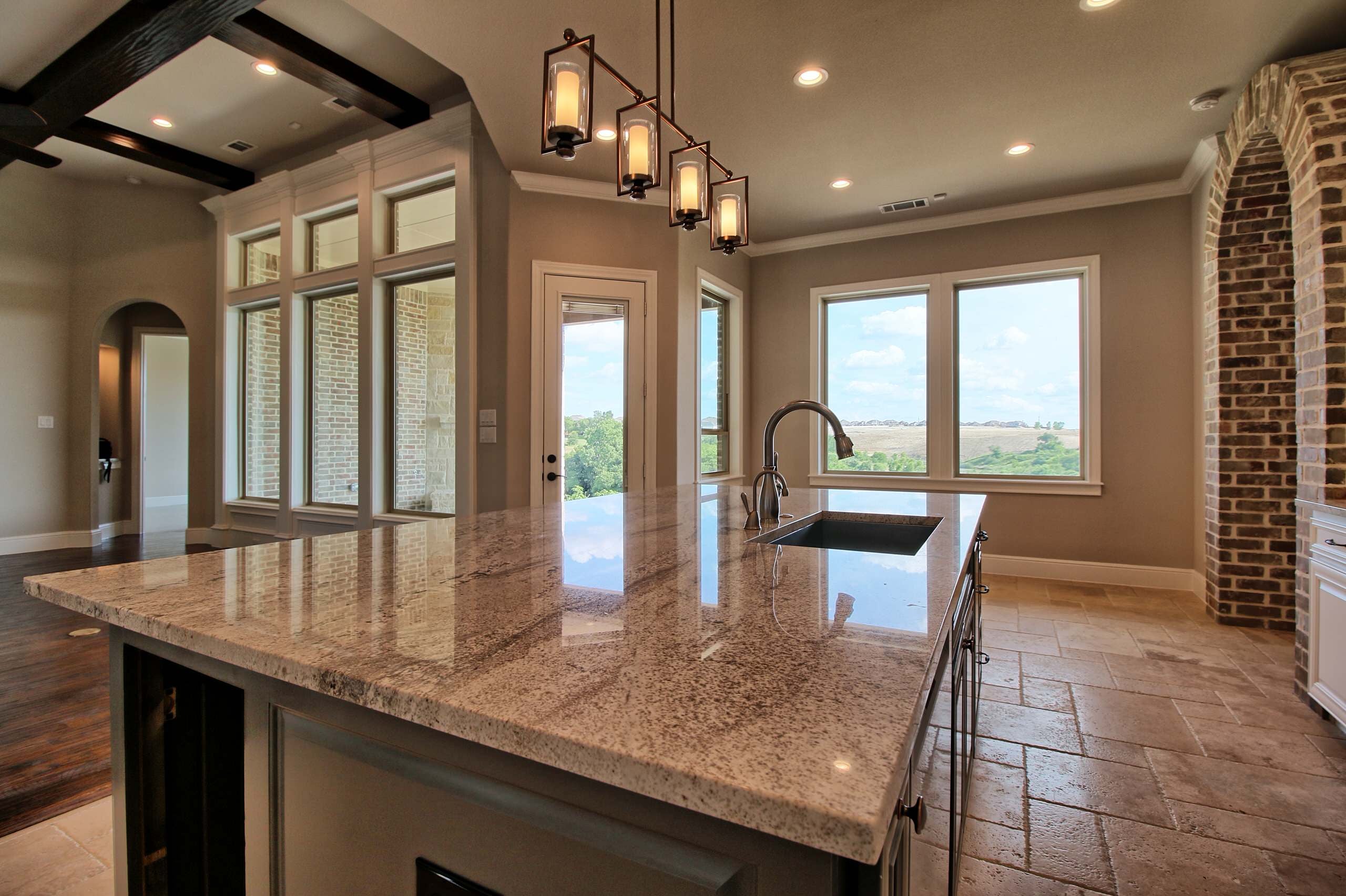 Granite kitchen island under a linear lantern pendant, with windows overlooking a hillside, a brick archway, and travertine floors