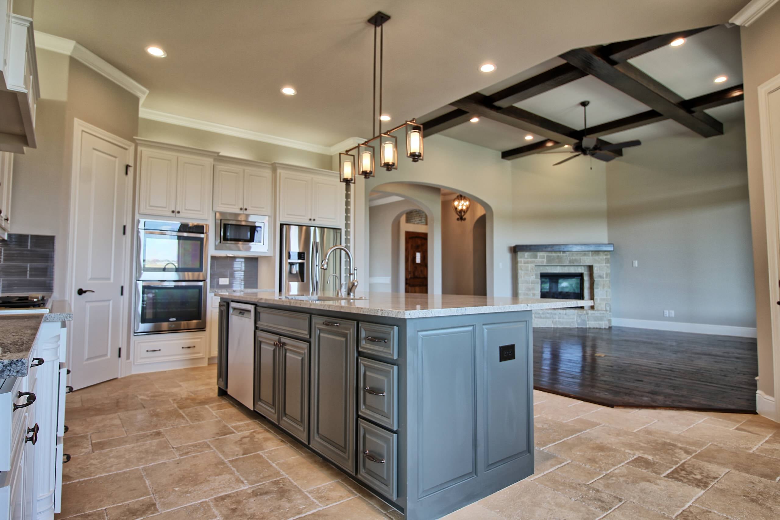 Kitchen with gray island, white cabinets, and stainless appliances, open to a living room with dark beamed ceiling and stone fireplace