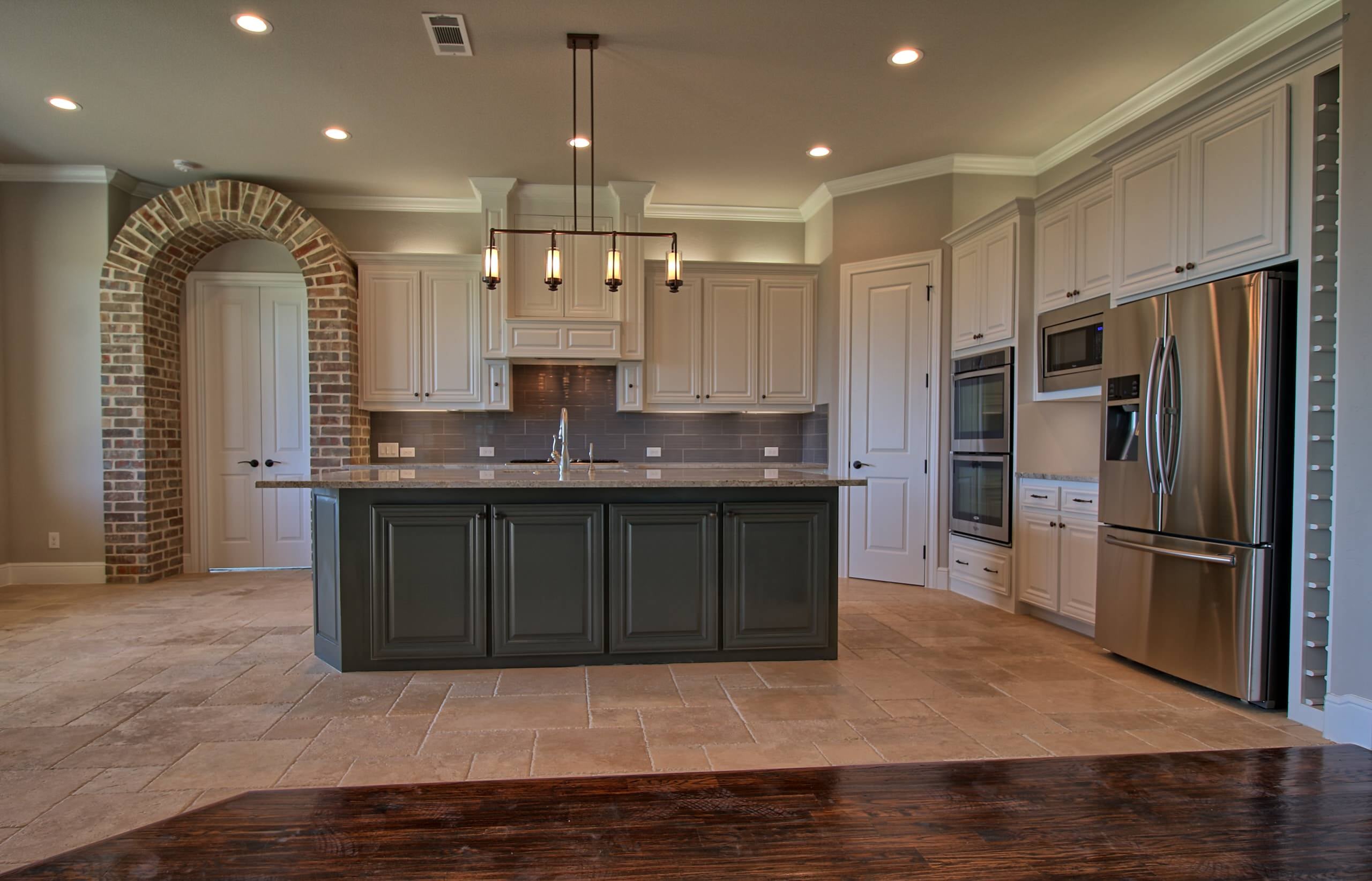 Kitchen with dark gray island, white cabinets, gray tile backsplash, stainless appliances, wine rack column, and a brick archway over double doors