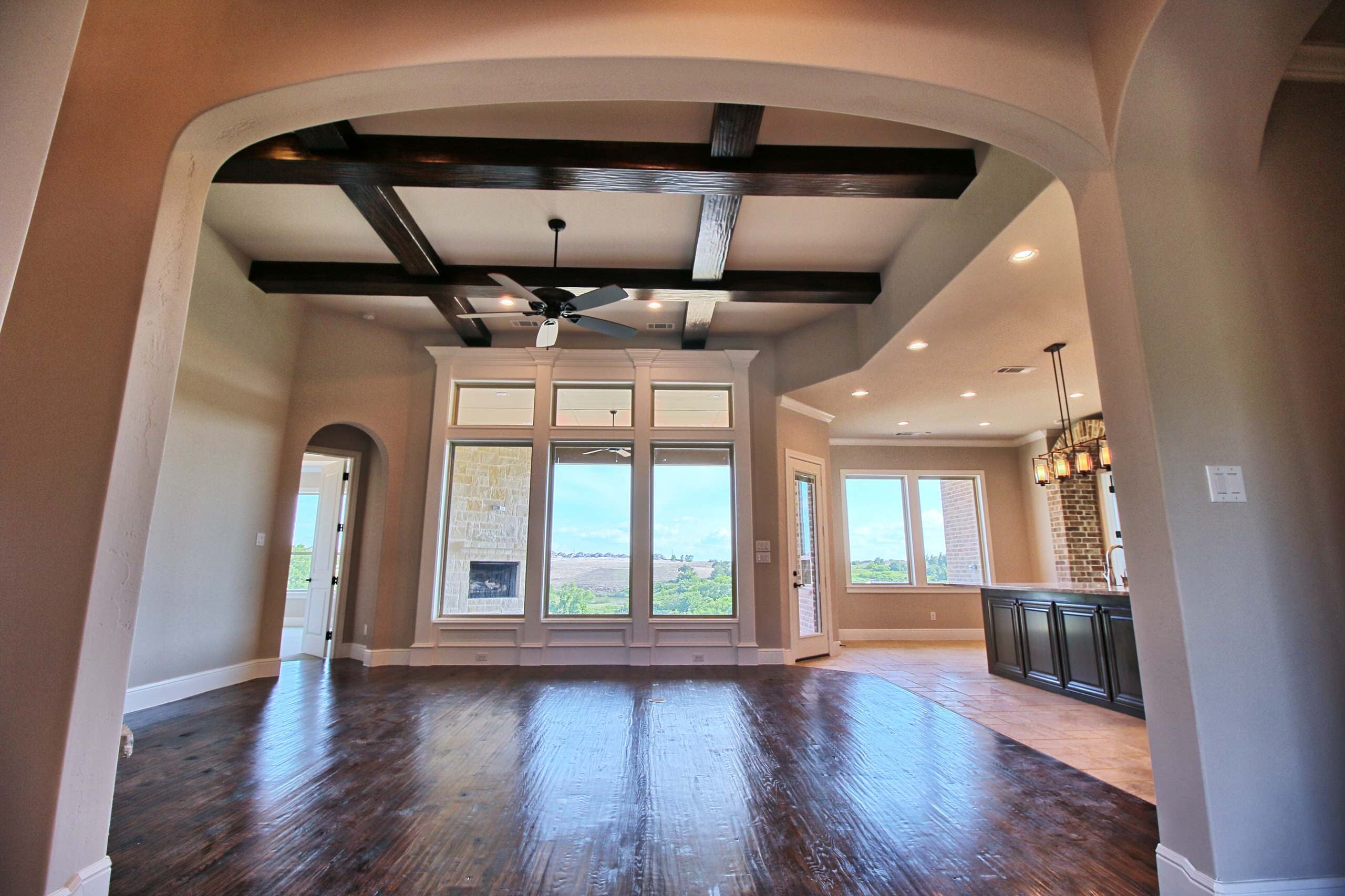 Great room framed by an arch, with dark wood ceiling beams, hand-scraped hardwood, and tall windows facing a covered patio and hills