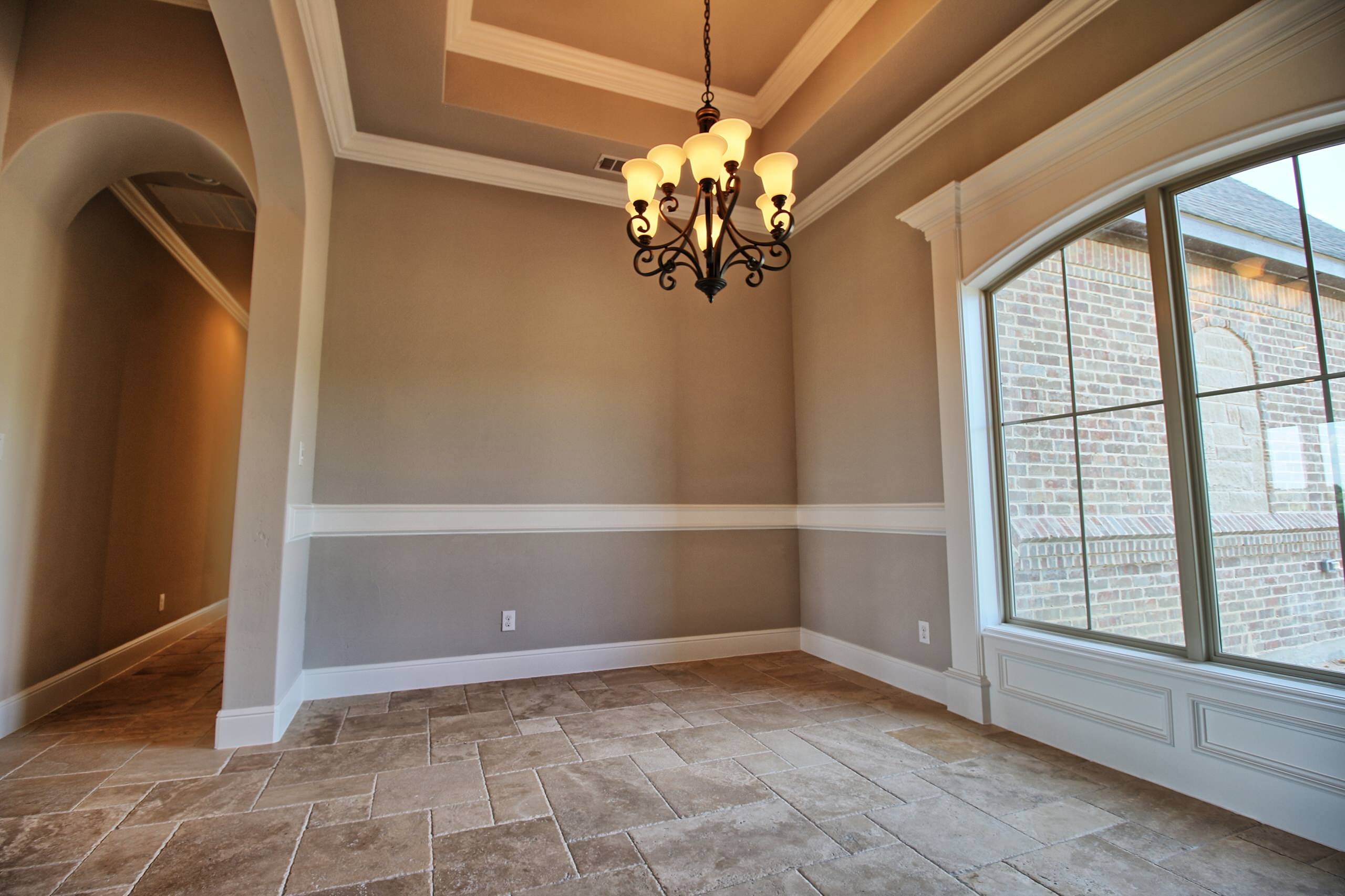 Dining room with tray ceiling, scrolled bronze chandelier, chair rail over two-tone gray walls, arched-top window, and travertine floors