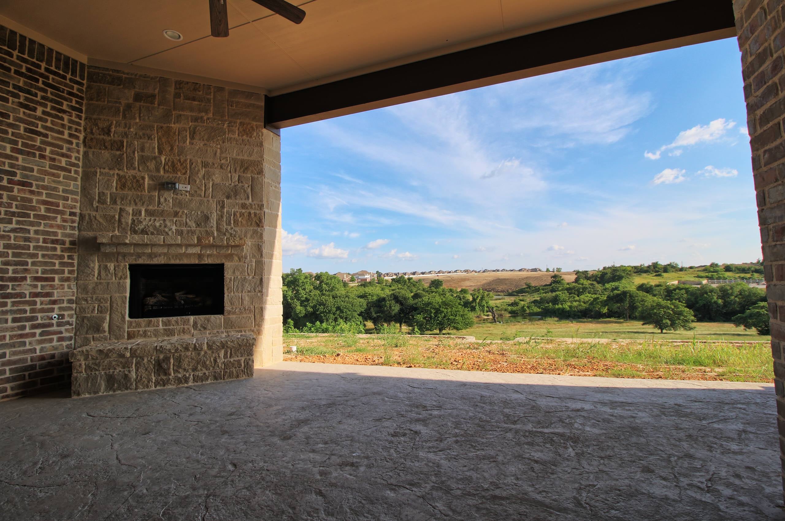 Covered patio with stamped concrete floor, cut-stone fireplace with raised hearth, brick walls, and ceiling fan, framing a view of wooded hills