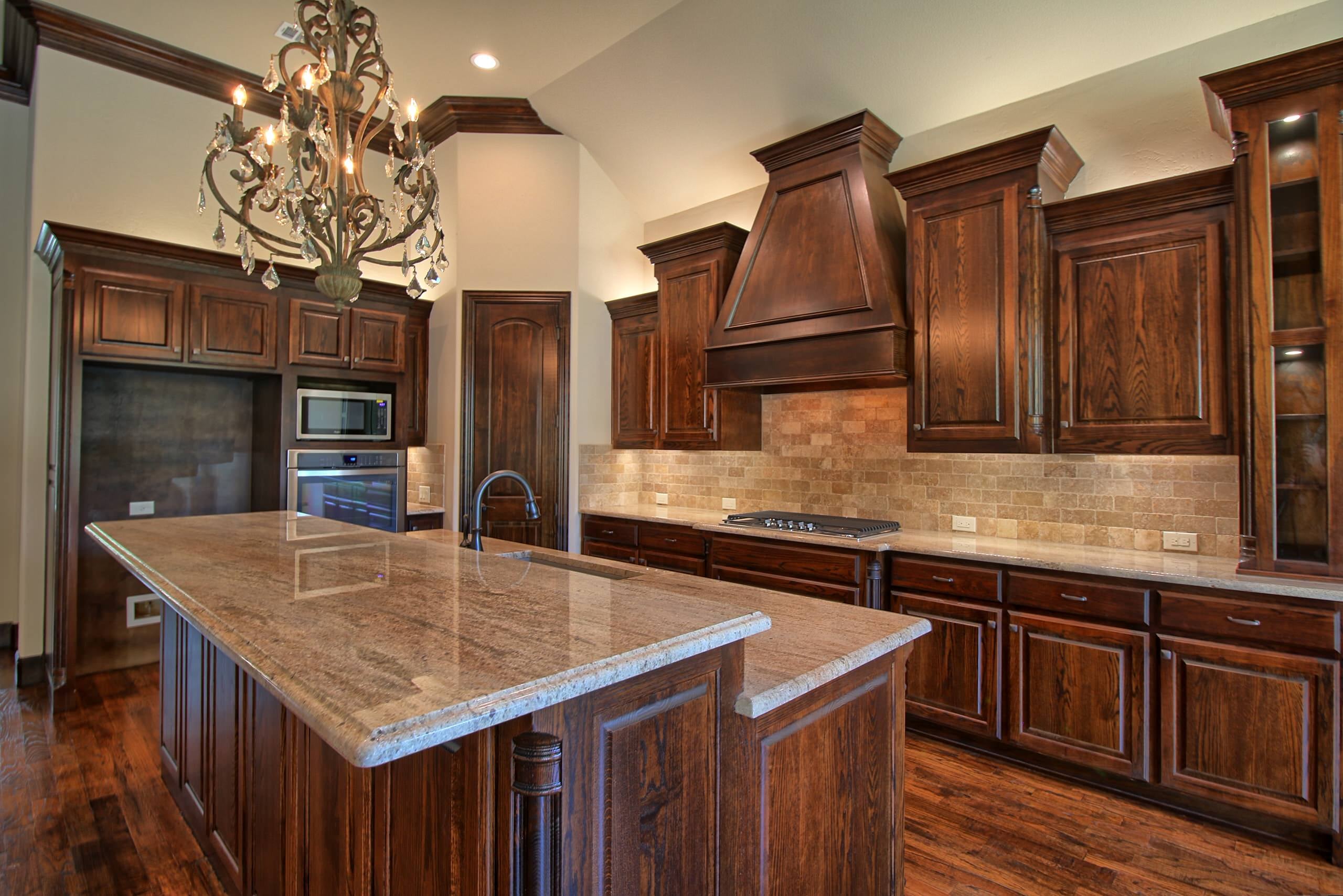 Kitchen with dark stained oak cabinetry, wood range hood, travertine subway backsplash, granite island with sink, crystal chandelier and hardwood floors