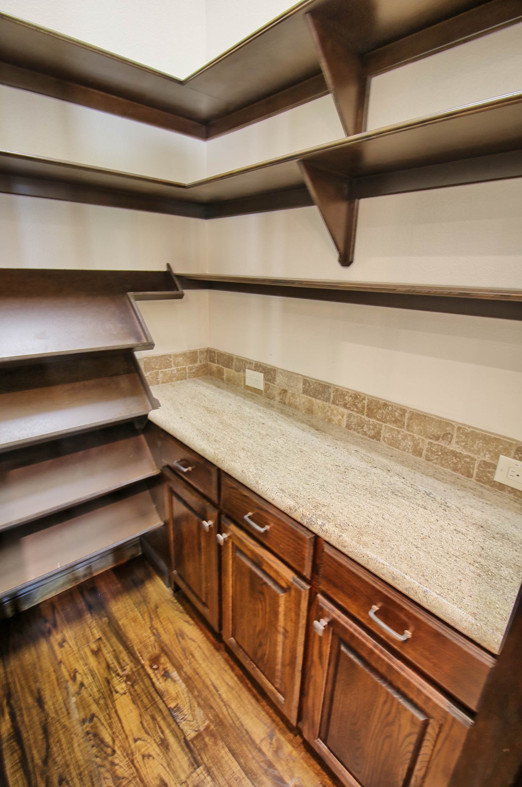 Walk-in pantry with dark stained wood shelving, angled shelves, oak base cabinets, granite countertop, travertine backsplash and hand-scraped hardwood floor