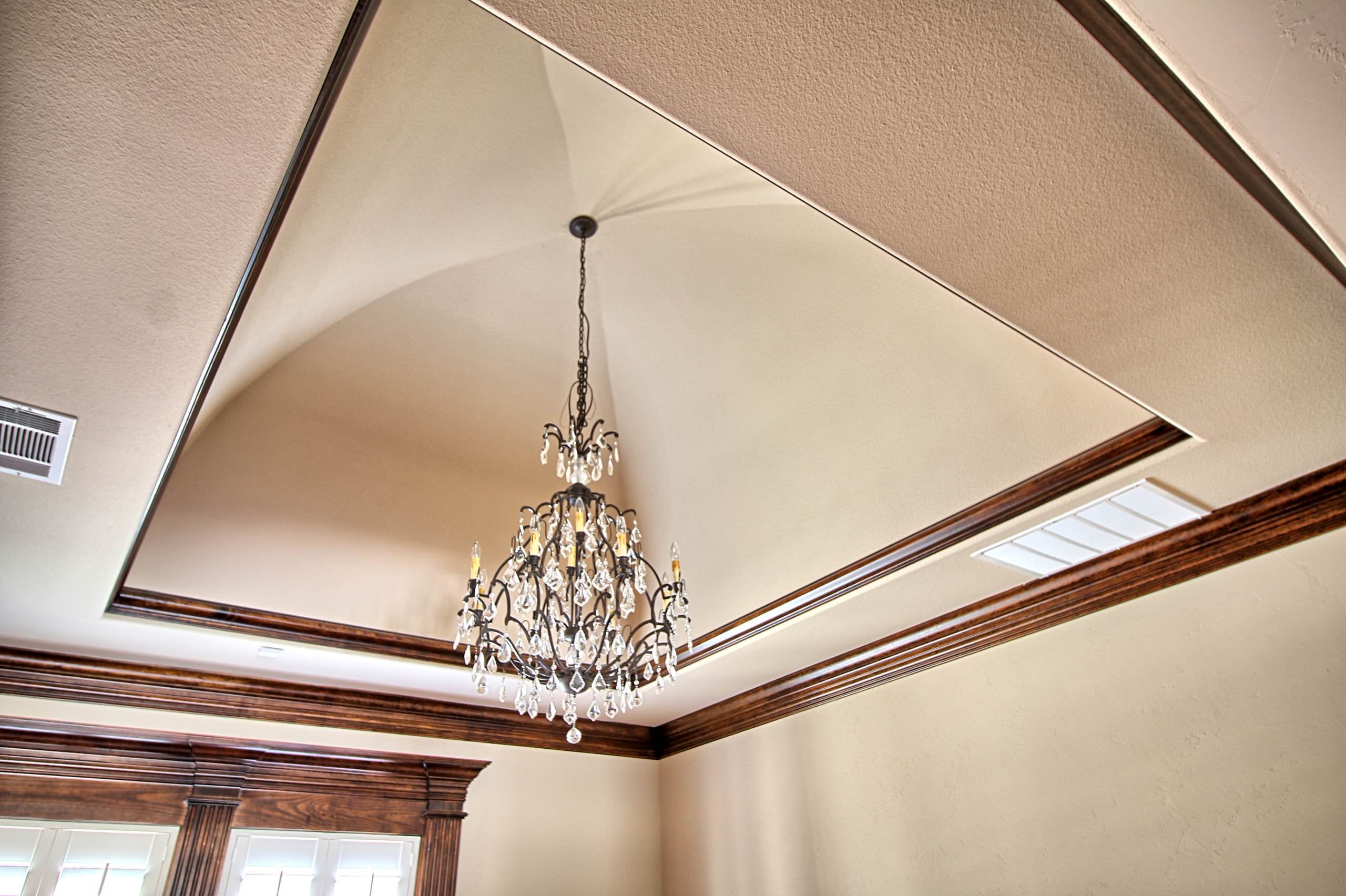 Groin-vaulted tray ceiling framed in stained wood crown molding, with a dark iron and crystal chandelier above shuttered windows