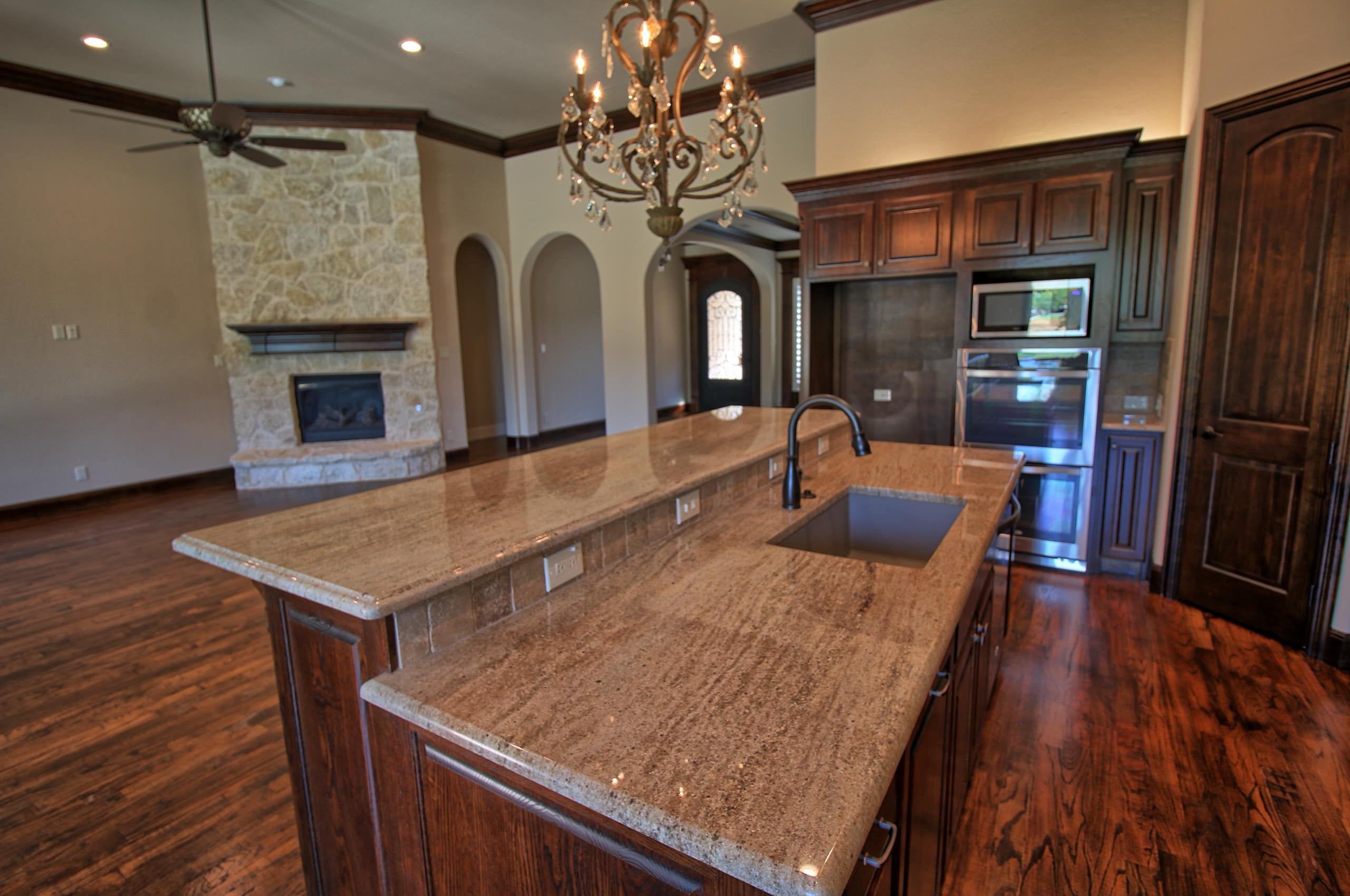 Two-level granite island with undermount sink under a crystal chandelier, open to a living area with stone fireplace and arched doorways