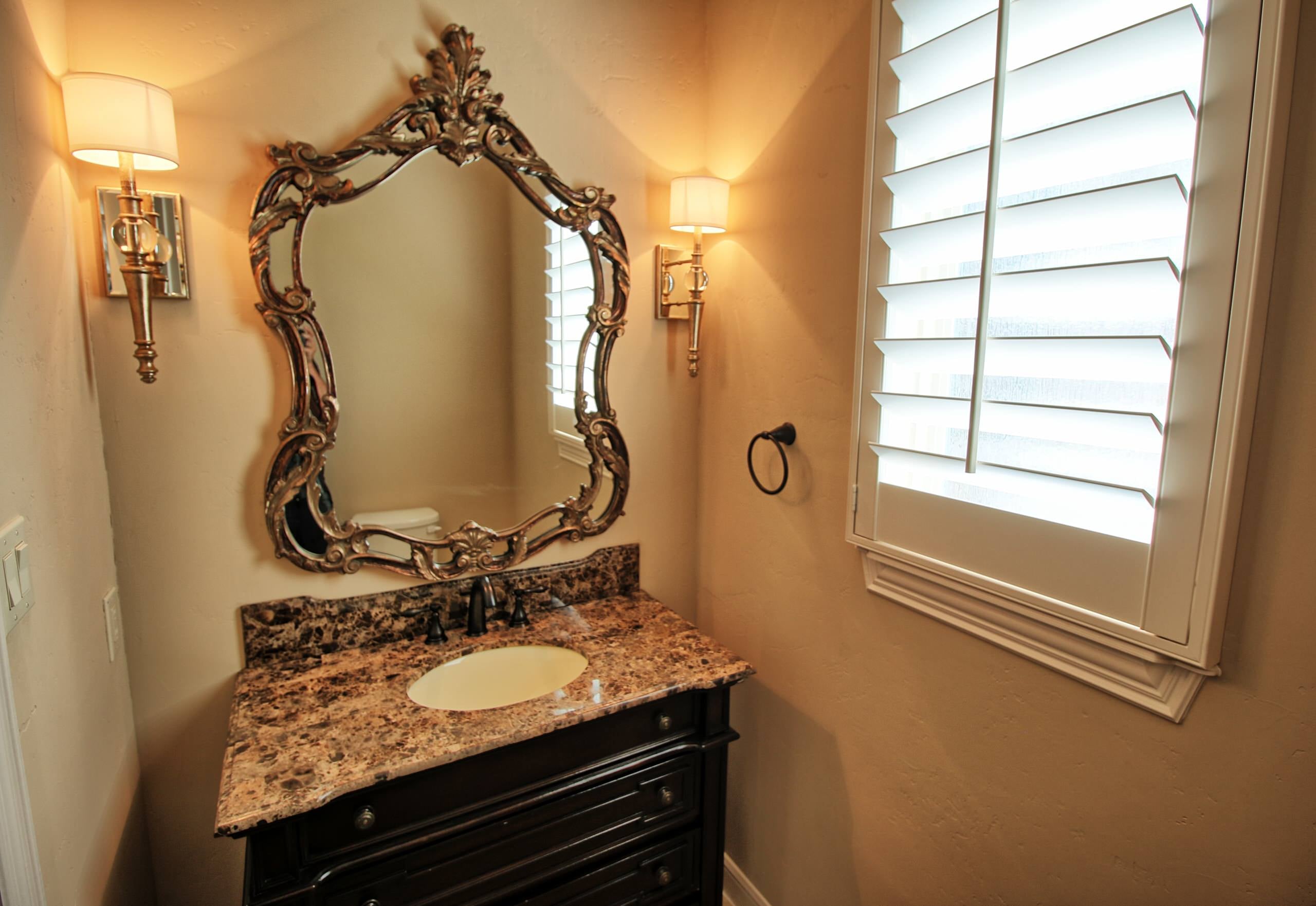 Powder room with dark furniture-style vanity, brown marble top, ornate gold-finish mirror, gold drum-shade sconces and white plantation shutters