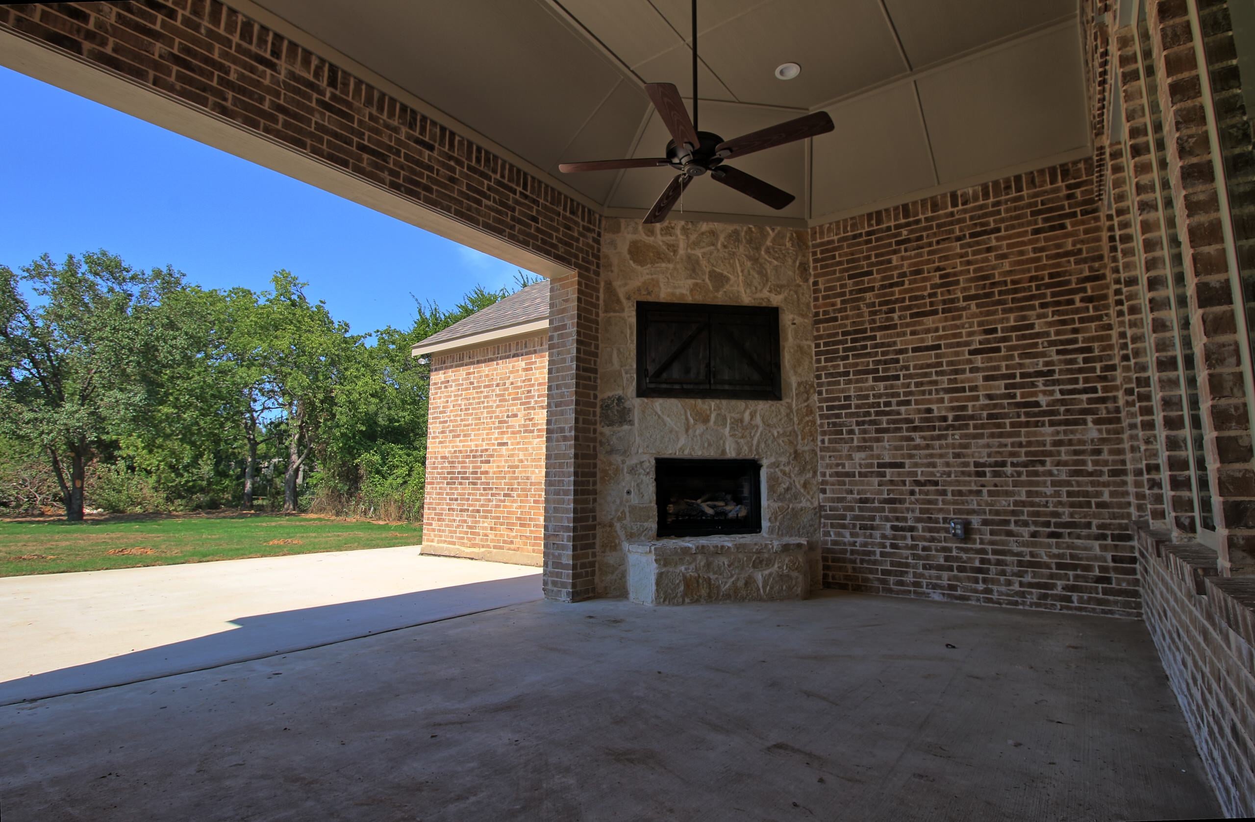 Covered patio with brick walls, stone corner fireplace with barn-door cabinet above, ceiling fan and concrete floor opening to a lawn