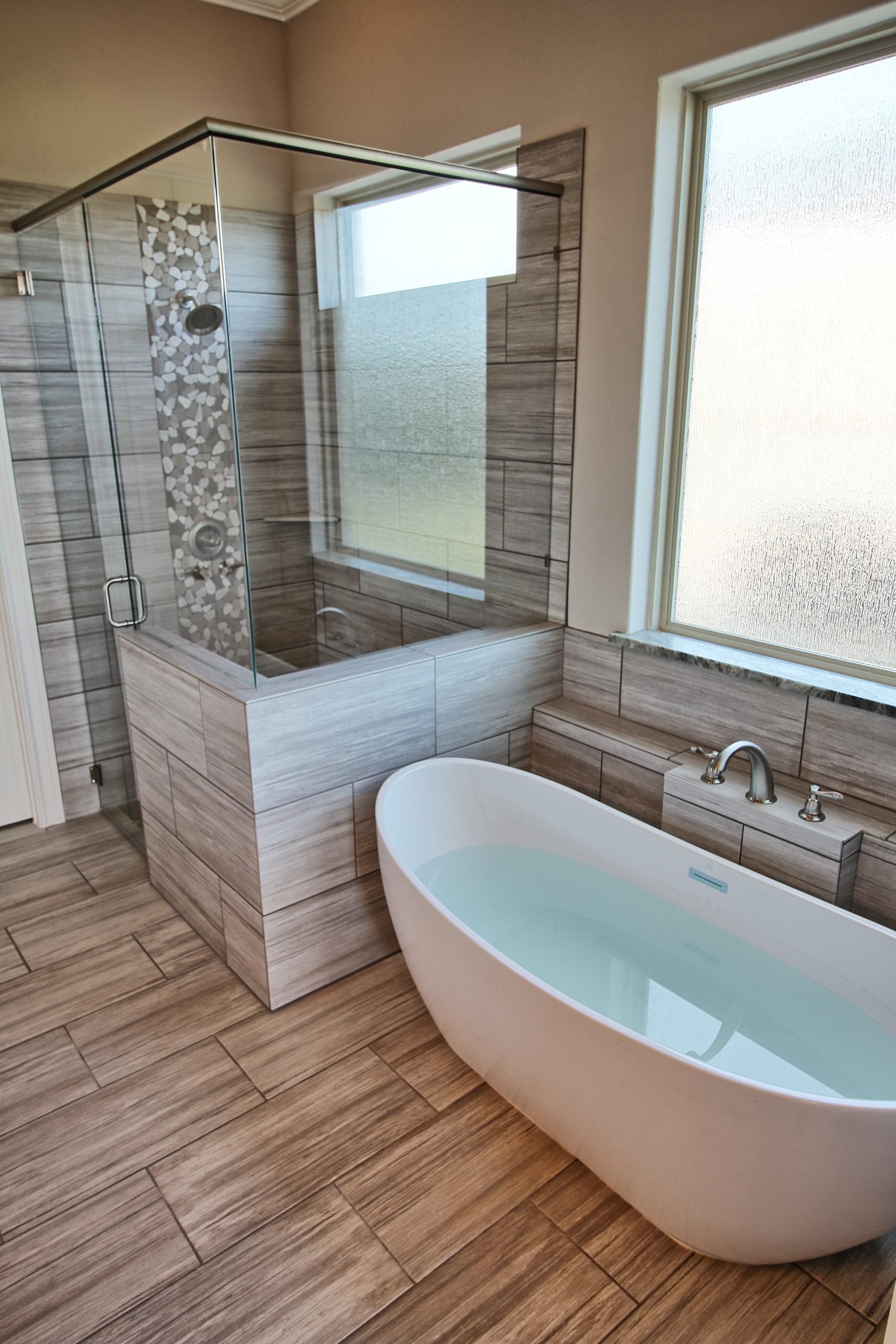 Freestanding soaking tub and glass corner shower with gray striated tile and pebble mosaic accent, textured glass windows and wood-look tile floor