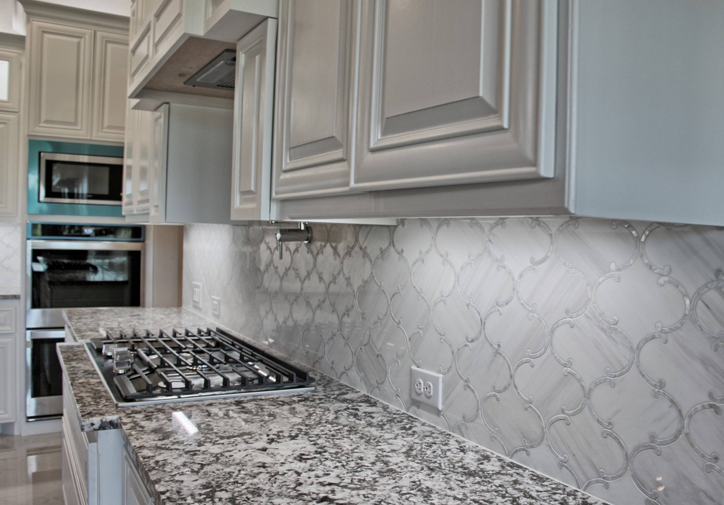Close view of a granite countertop with stainless gas cooktop, arabesque marble mosaic backsplash, pot filler and white raised-panel cabinets