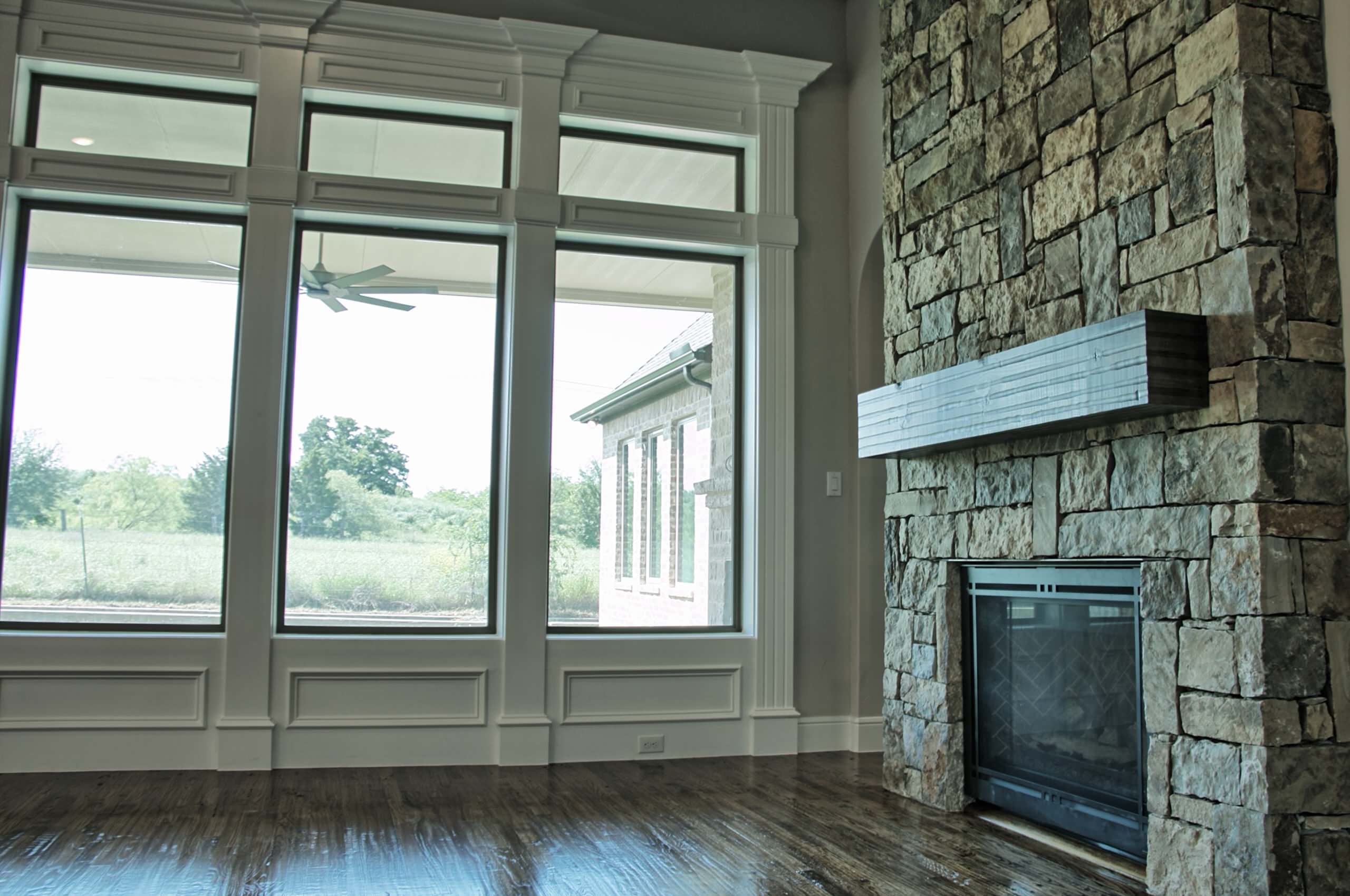 Living room with stacked stone fireplace, rough-sawn beam mantel, tall windows and transoms in white paneled millwork, and dark hardwood floors