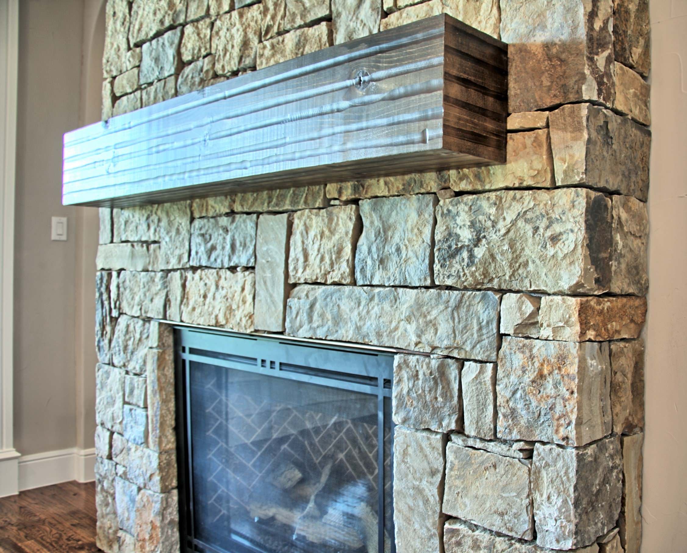 Close view of a stacked stone fireplace with a rough-sawn wood beam mantel and black metal screen over a herringbone-pattern firebox