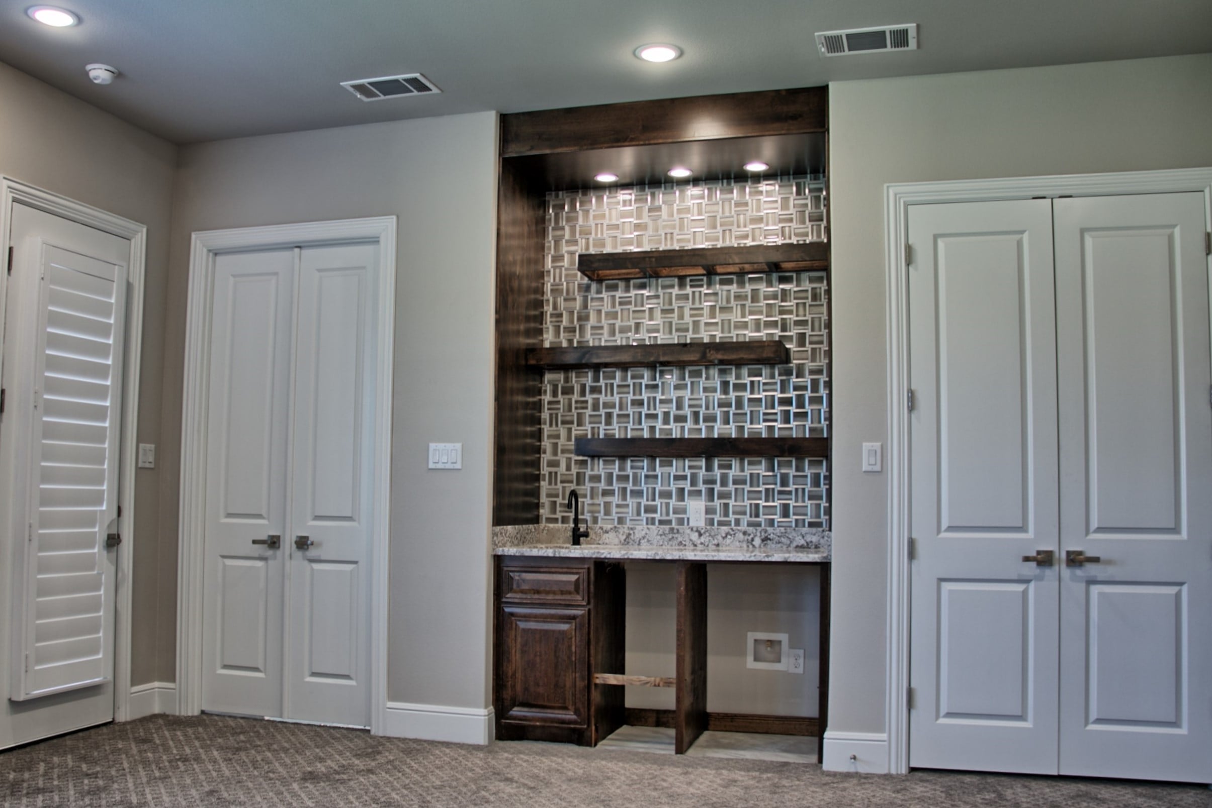 Built-in wet bar with dark stained wood surround, floating shelves, glass mosaic backsplash and granite counter, flanked by white double doors