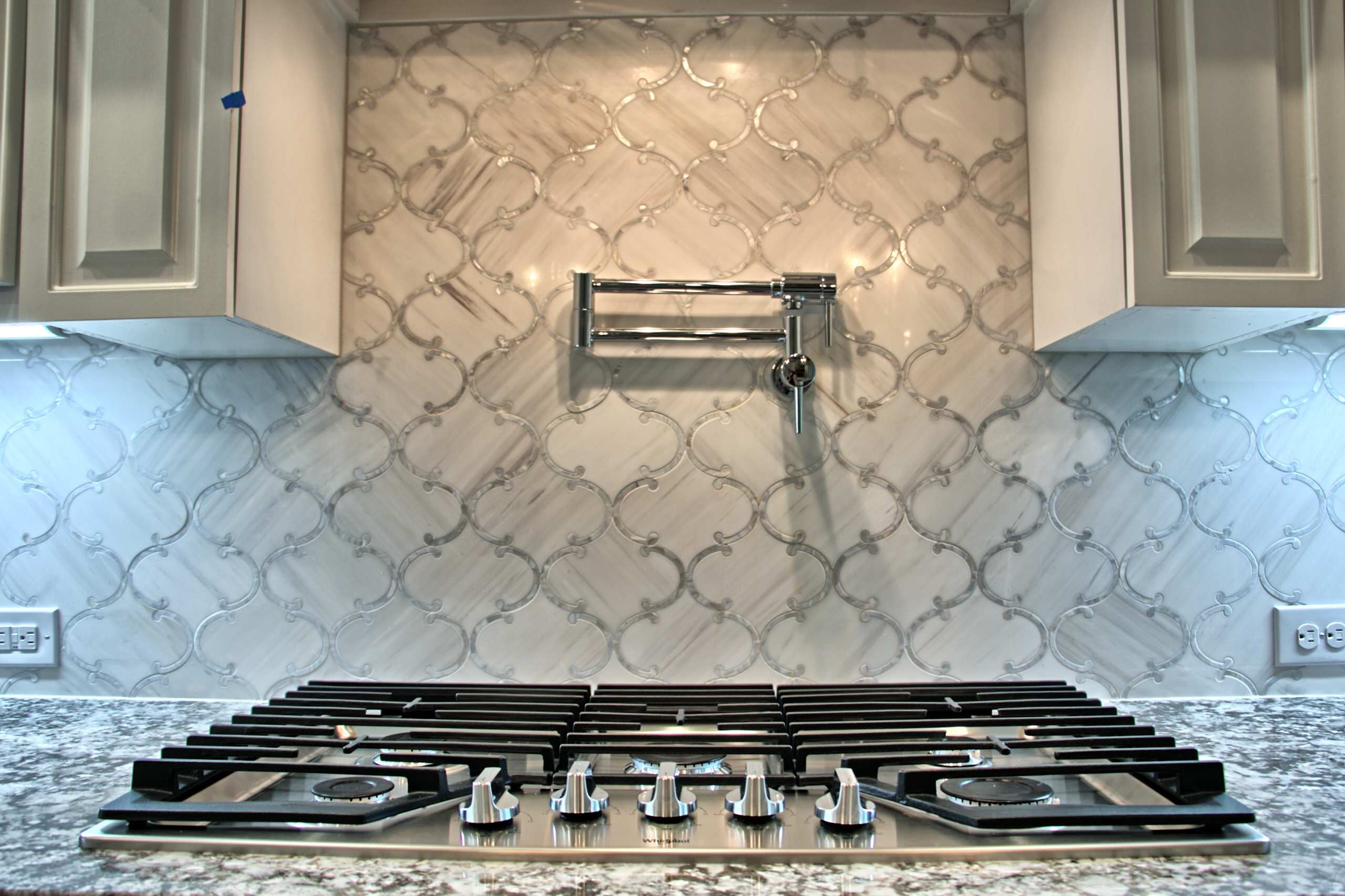 Stainless five-burner gas cooktop set in granite beneath a chrome pot filler and arabesque marble mosaic backsplash, framed by white raised-panel cabinets