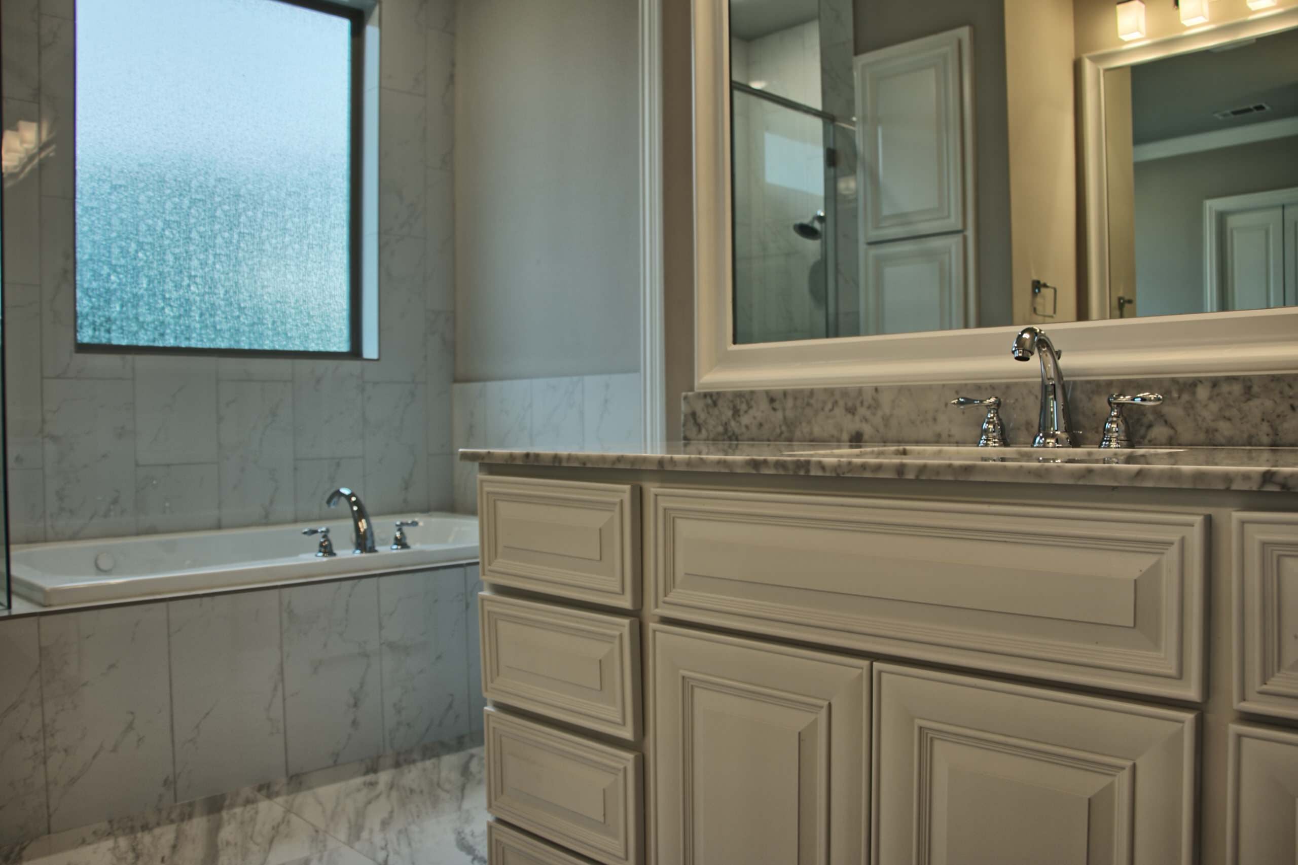 Drop-in tub with marble-look tile surround under a textured glass window, beside a white raised-panel vanity with marble top and framed mirror