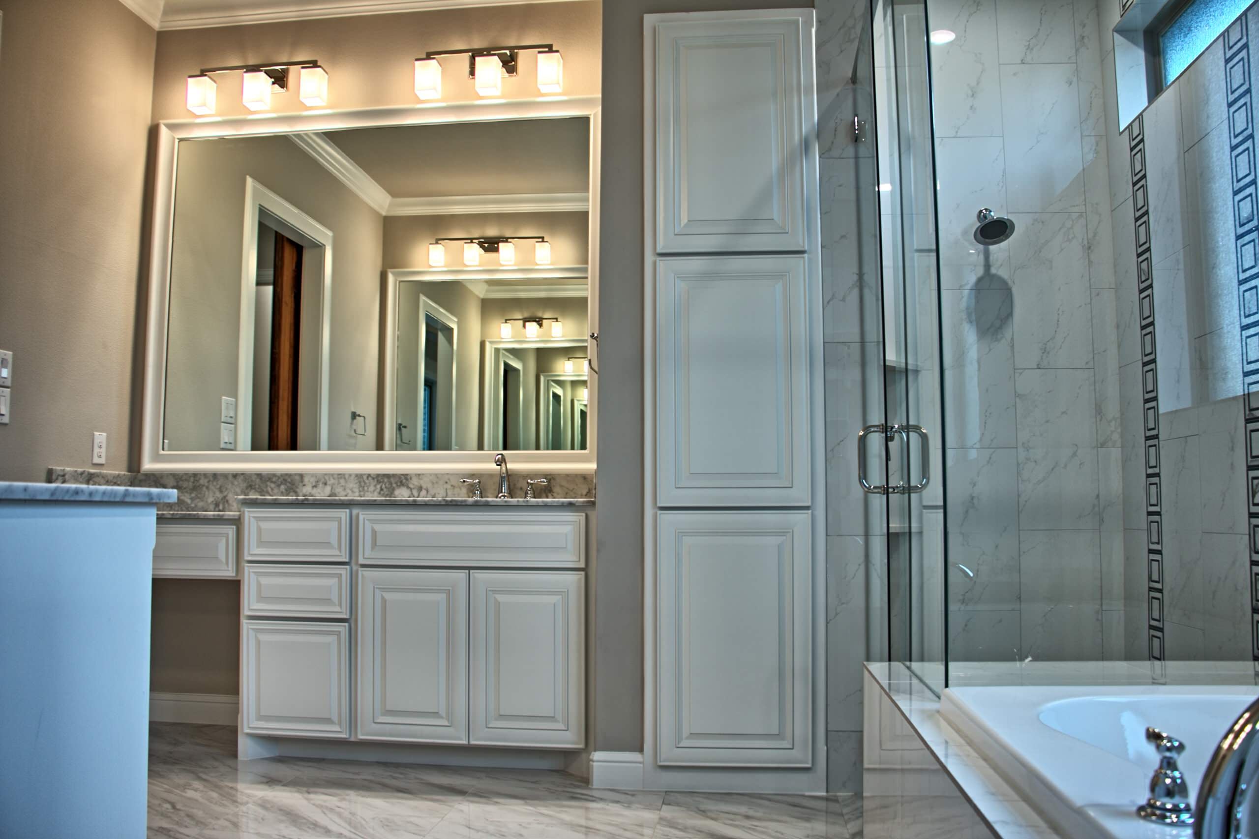 White raised-panel vanity with marble top, framed mirror and frosted cube vanity lights, beside a tall linen cabinet and frameless glass shower