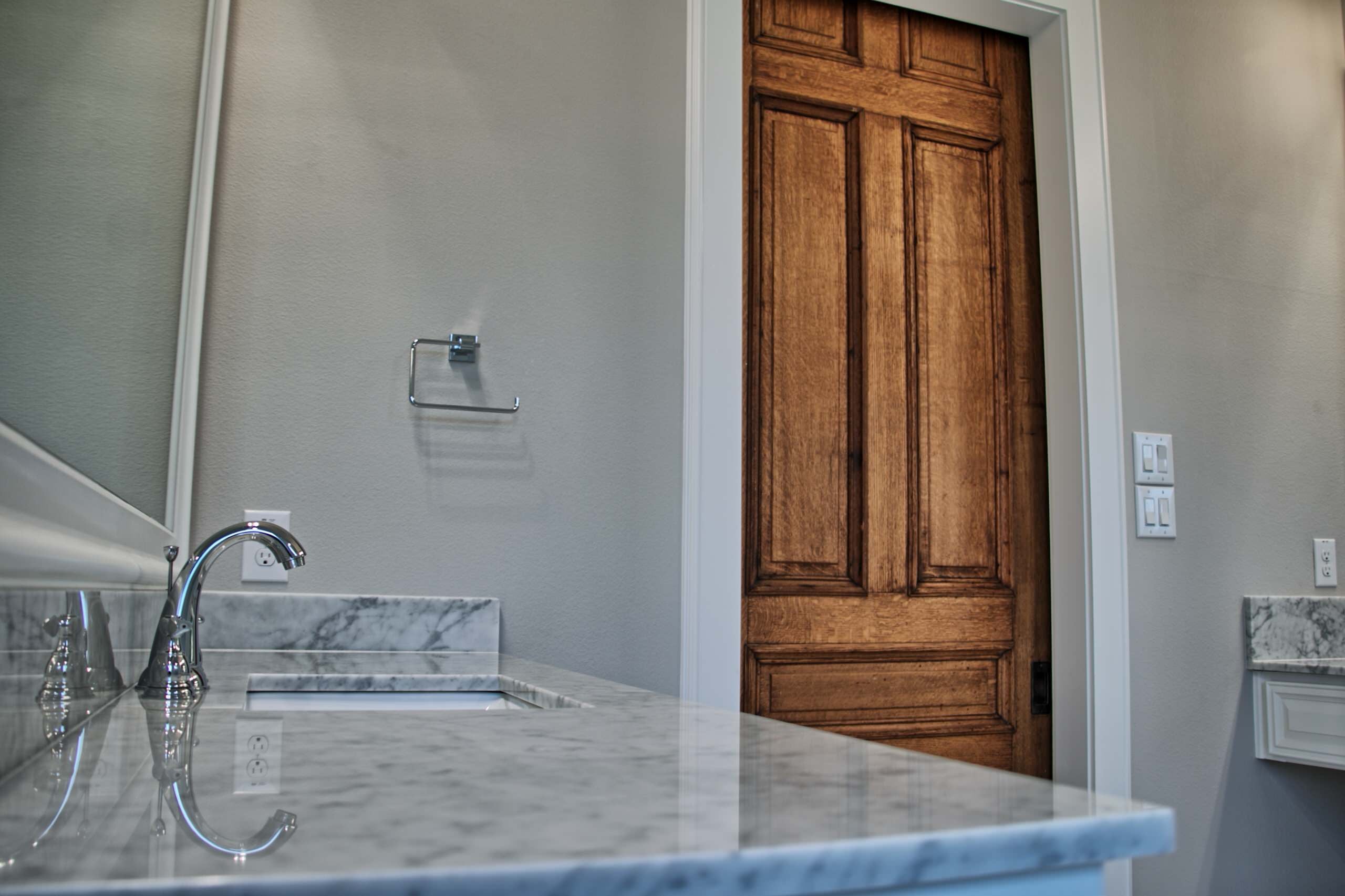 Marble vanity top with undermount sink and chrome faucet in the foreground, with a stained oak paneled door in white casing beyond
