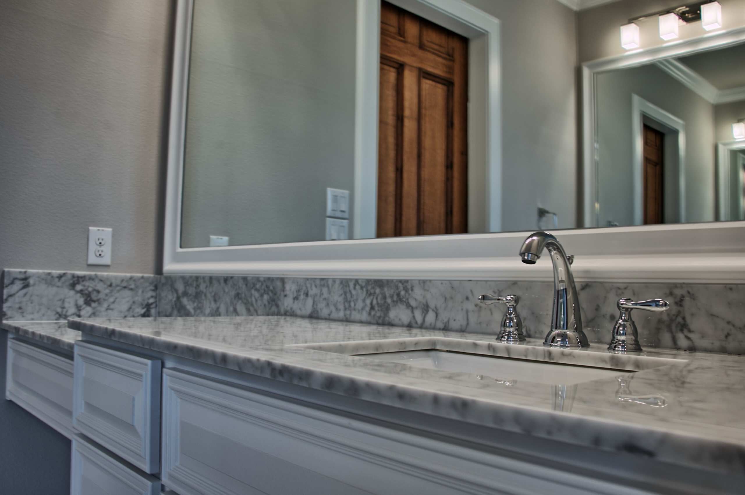 White raised-panel vanity with marble countertop, undermount sink and chrome widespread faucet beneath a white-framed mirror reflecting a wood door