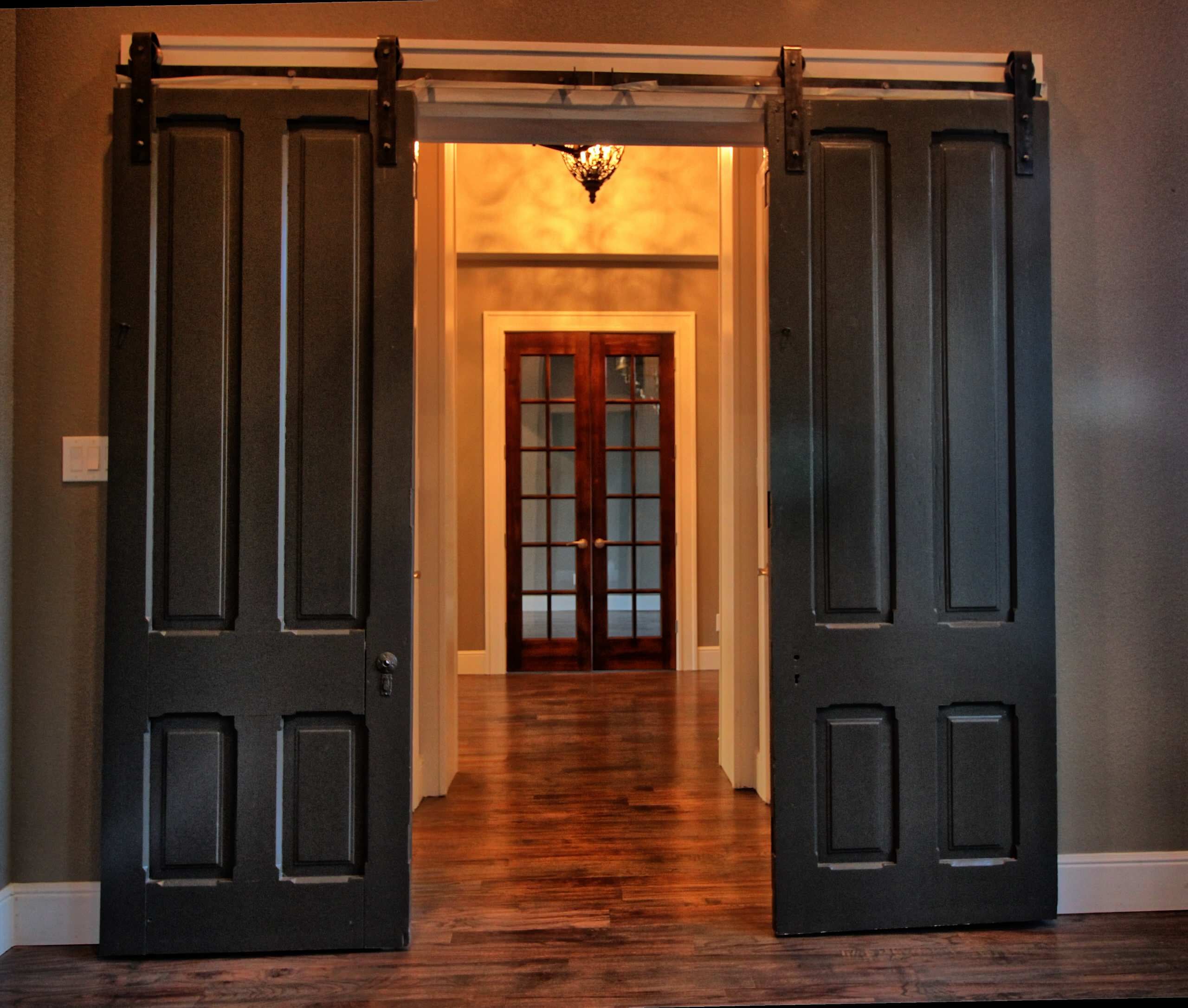 Pair of dark painted antique-style panel doors on sliding barn door hardware, opening to a hardwood hallway and wood French doors