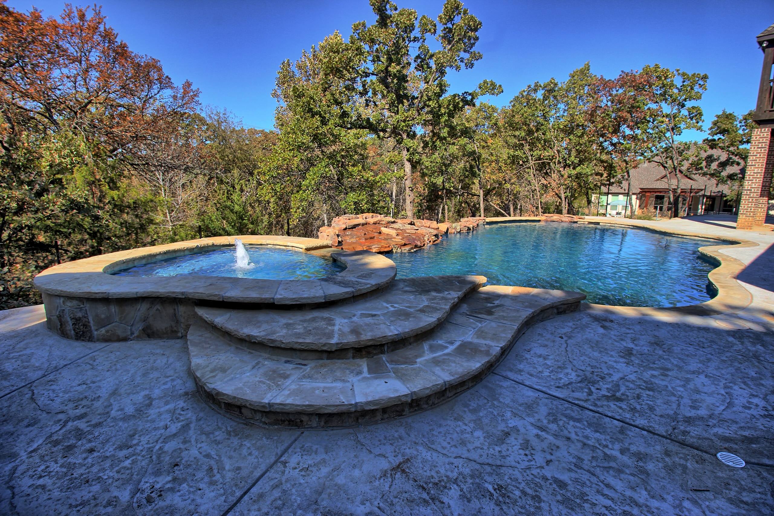 Raised stone spa with bubbler and curved flagstone steps beside a freeform pool with boulder waterfall, stamped concrete deck and wooded backdrop