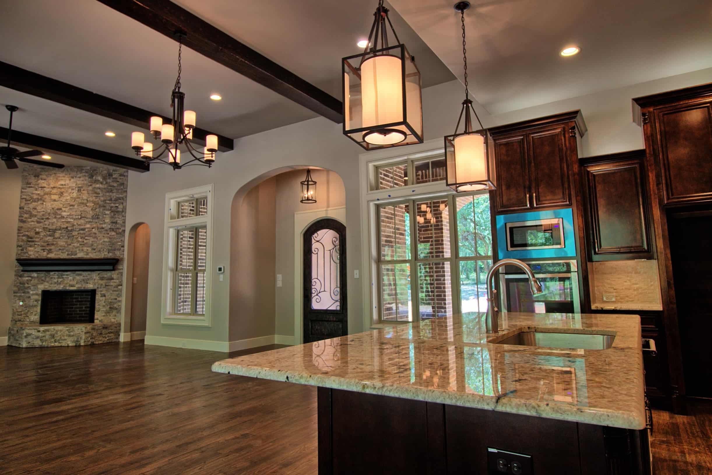 Kitchen island with granite top and lantern pendants beside dark wood cabinetry, facing a beamed living room with stacked stone corner fireplace and arched iron-detail door