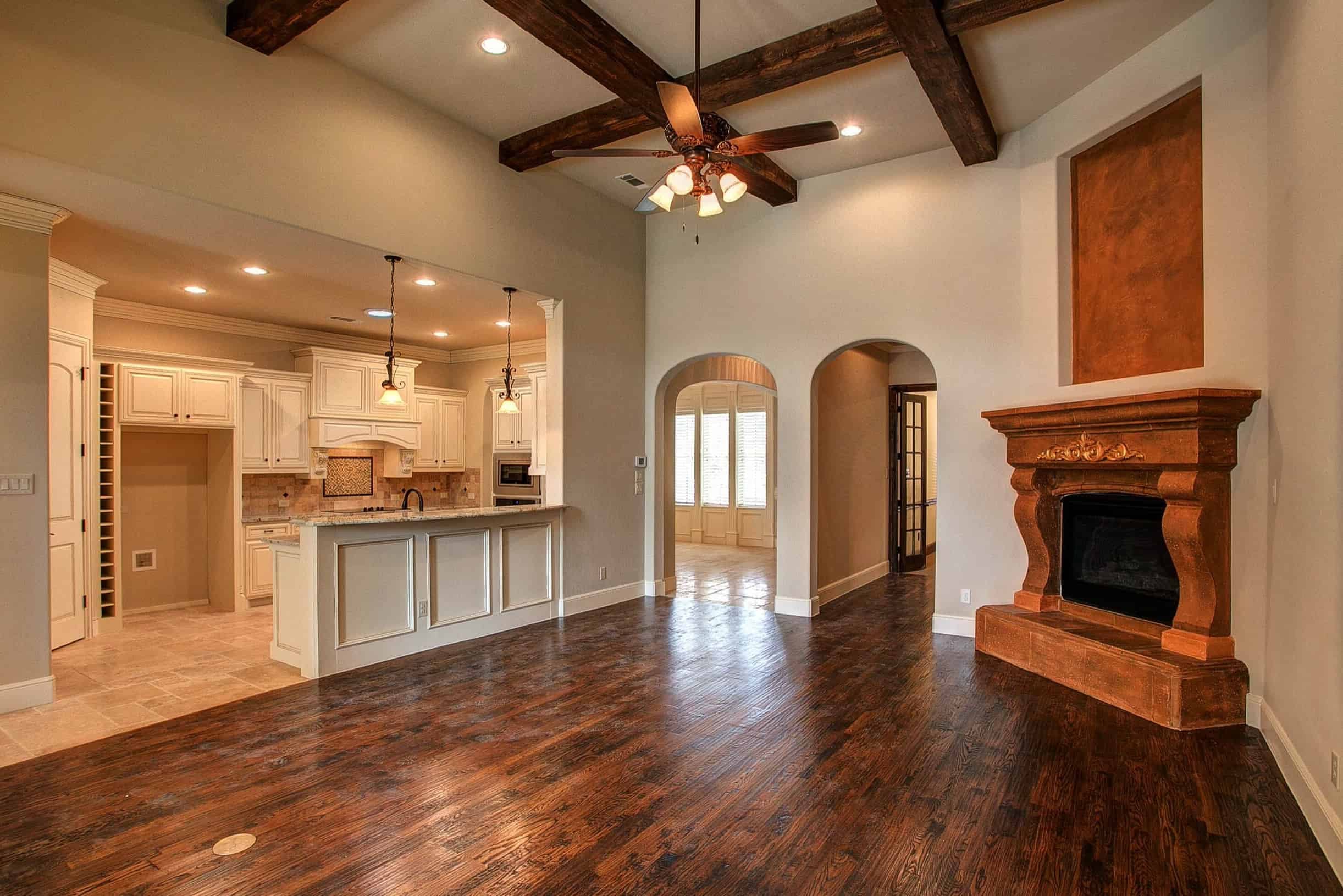 Living room with dark wood ceiling beams, hand-scraped wood floors and carved cast-stone corner fireplace, open to a cream kitchen with granite bar and arched doorways